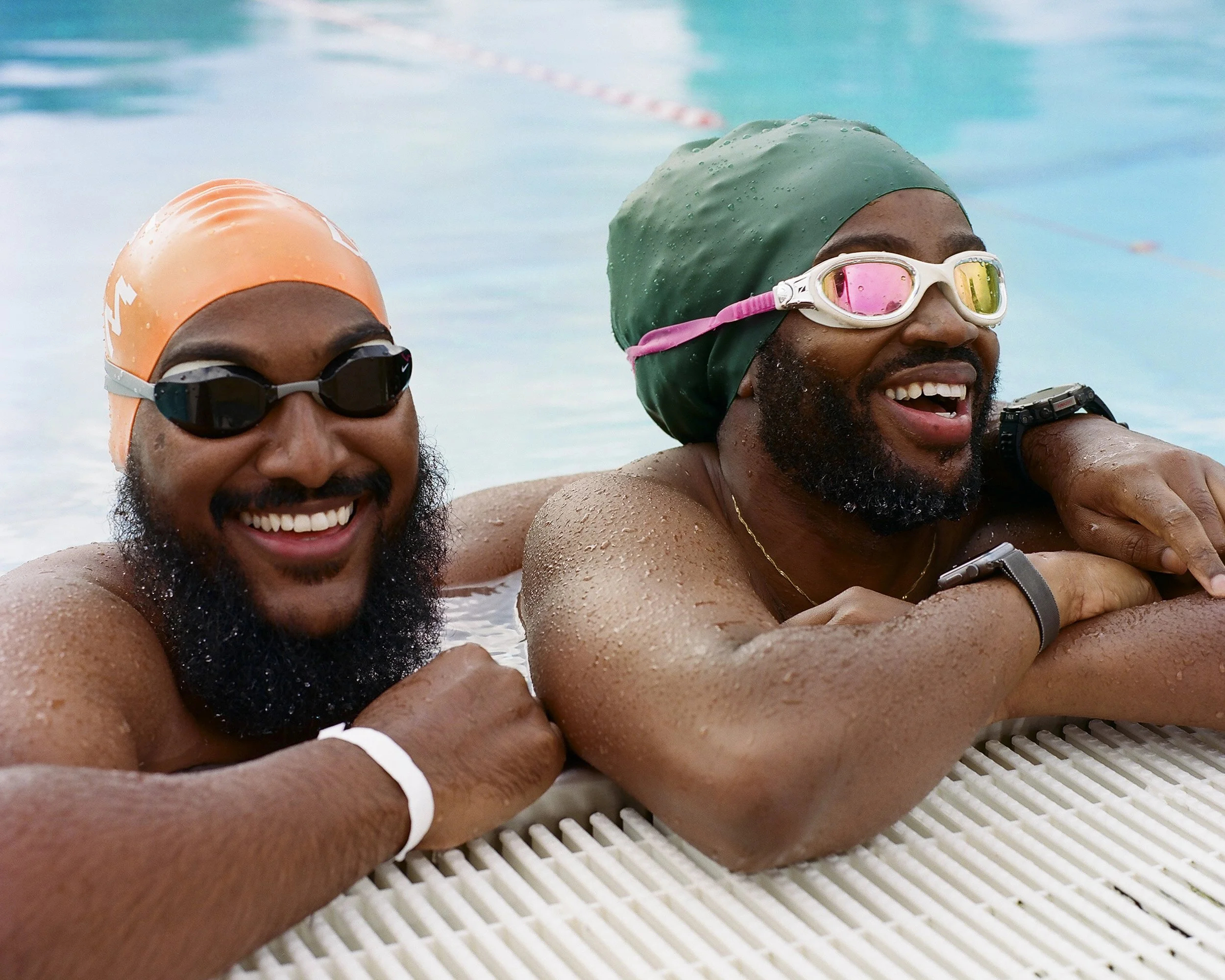 Two smiling men with beards wearing swimming goggles and caps lean on pool edge, turquoise water behind them.