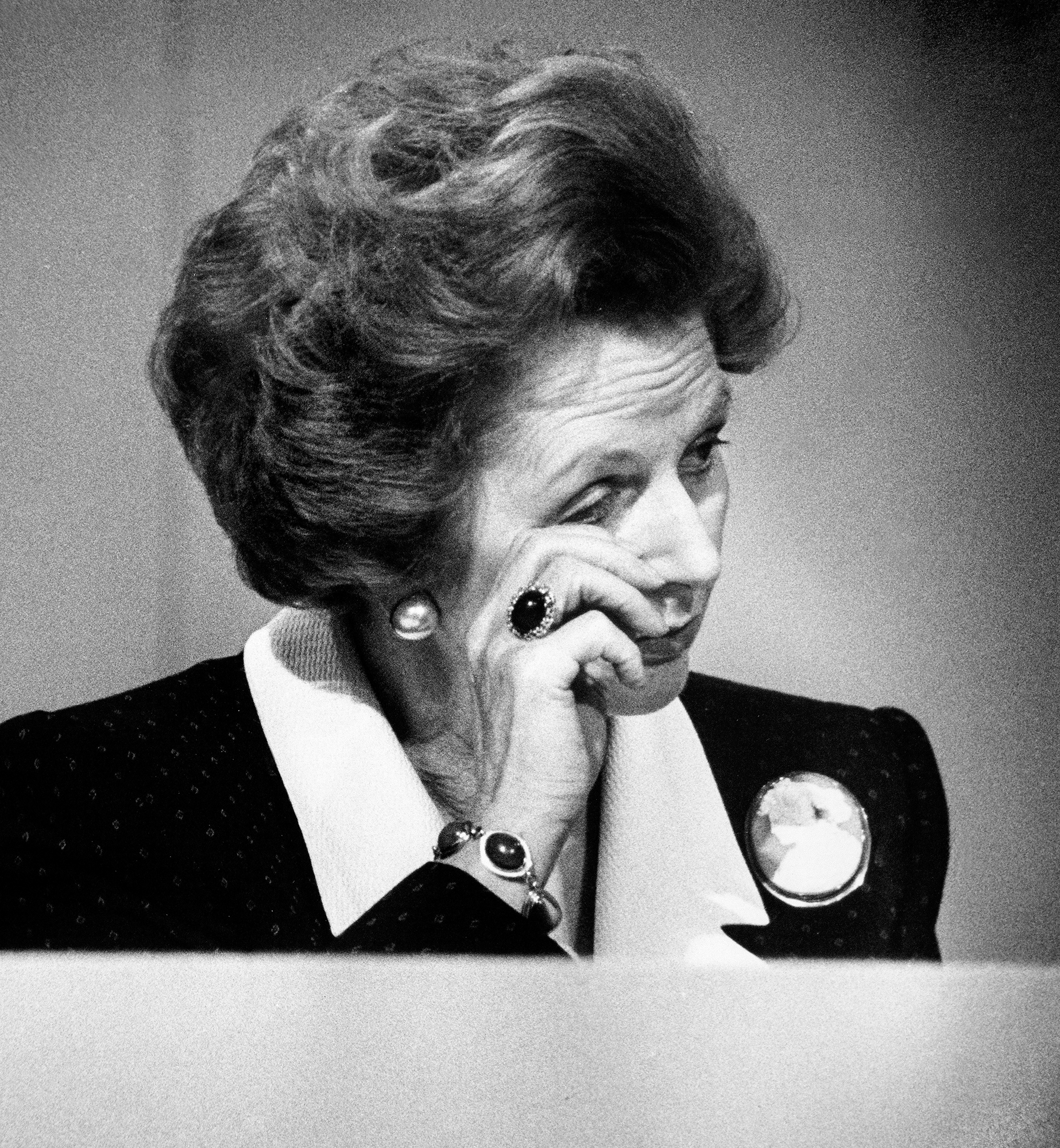 Black and white portrait of woman with voluminous hair, hand to chin, wearing dark jacket with white collar and circular brooch.