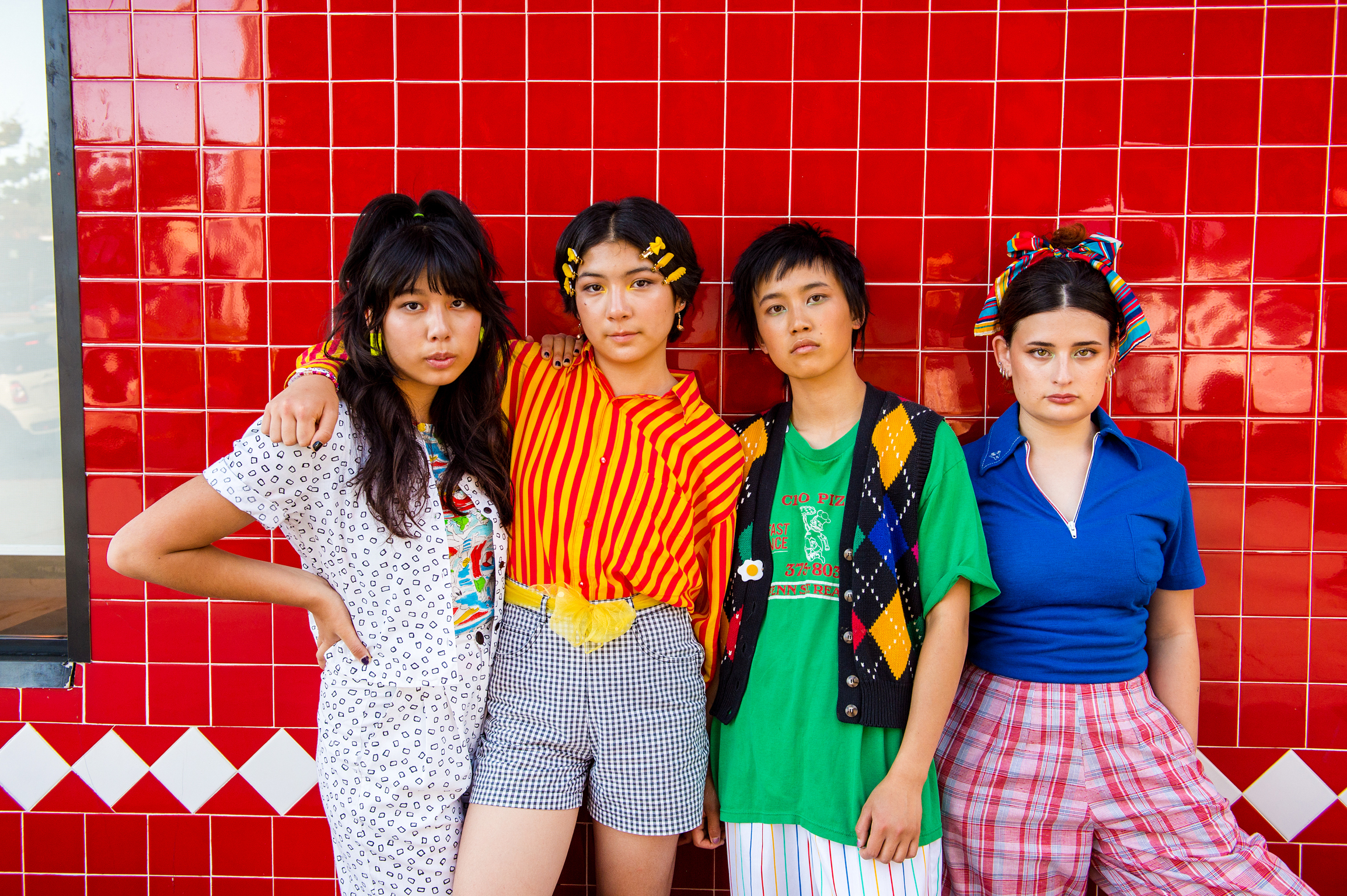 Four young women in colourful vintage-style outfits pose against bright red tiled wall with white diamond border pattern.