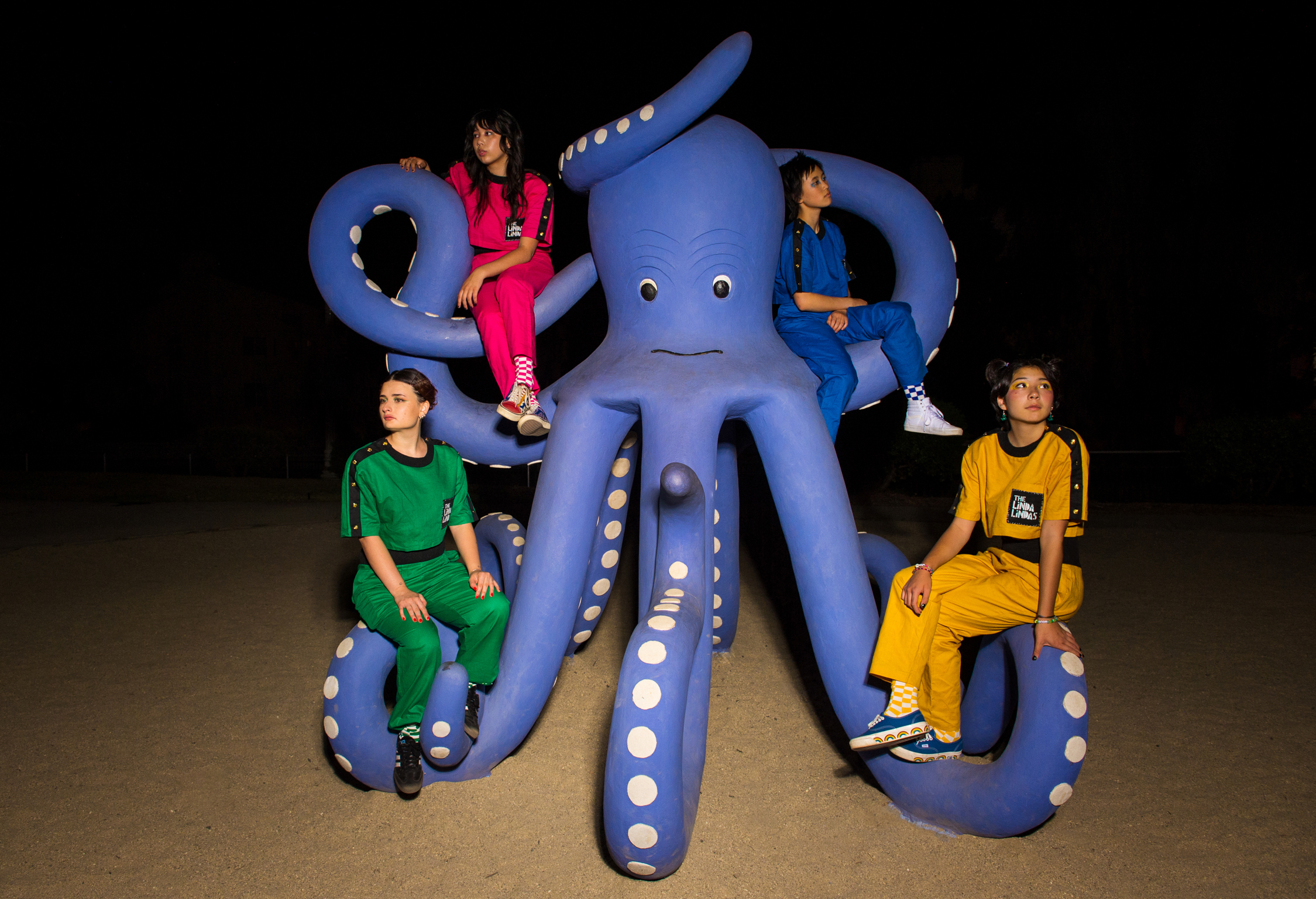 Four people sitting on large blue inflatable octopus with white spots on tentacles against black background. People wearing red, blue, green, and yellow clothing.