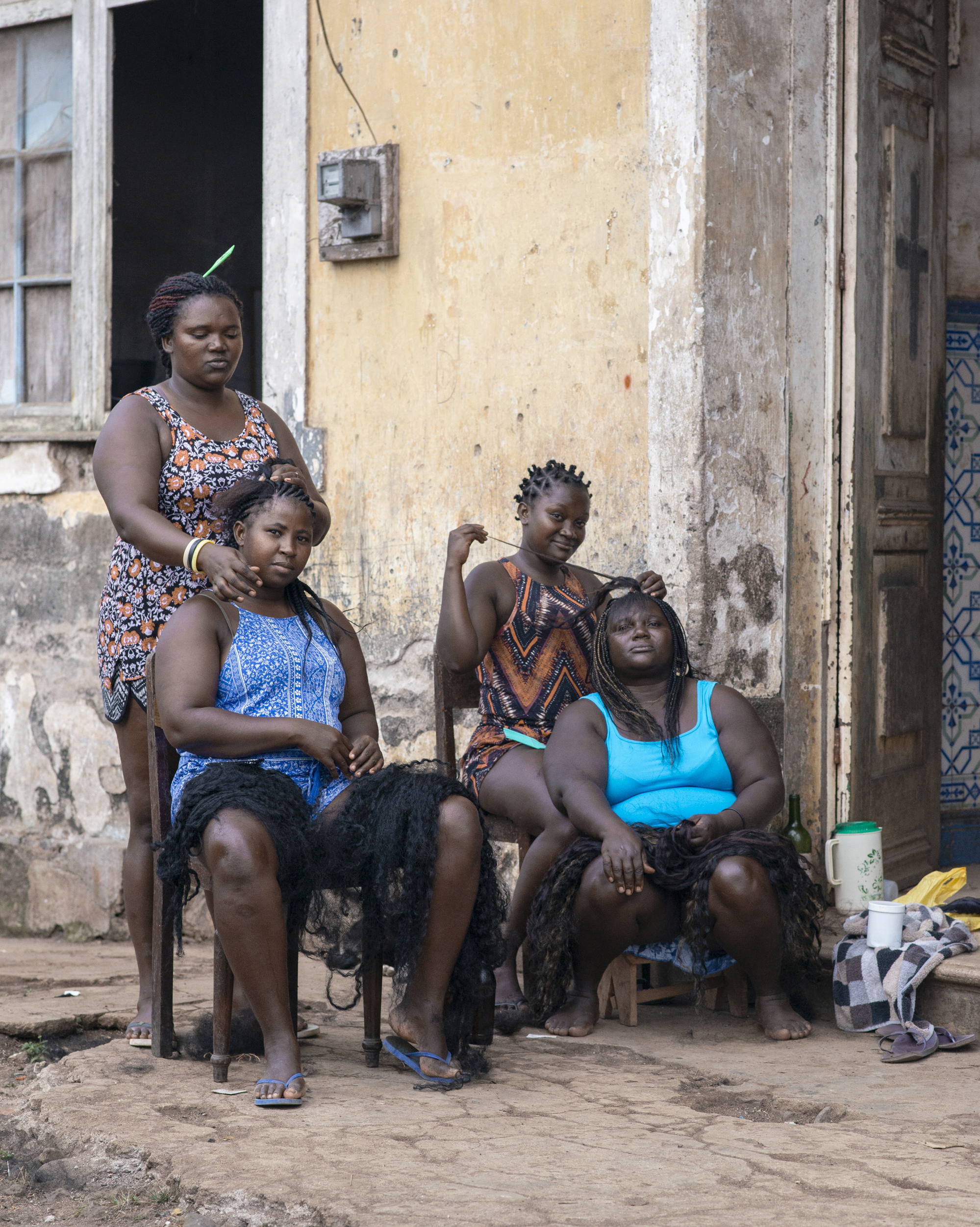 Woman standing behind seated woman having hair braided, with another woman braiding a child's hair, outside weathered building.