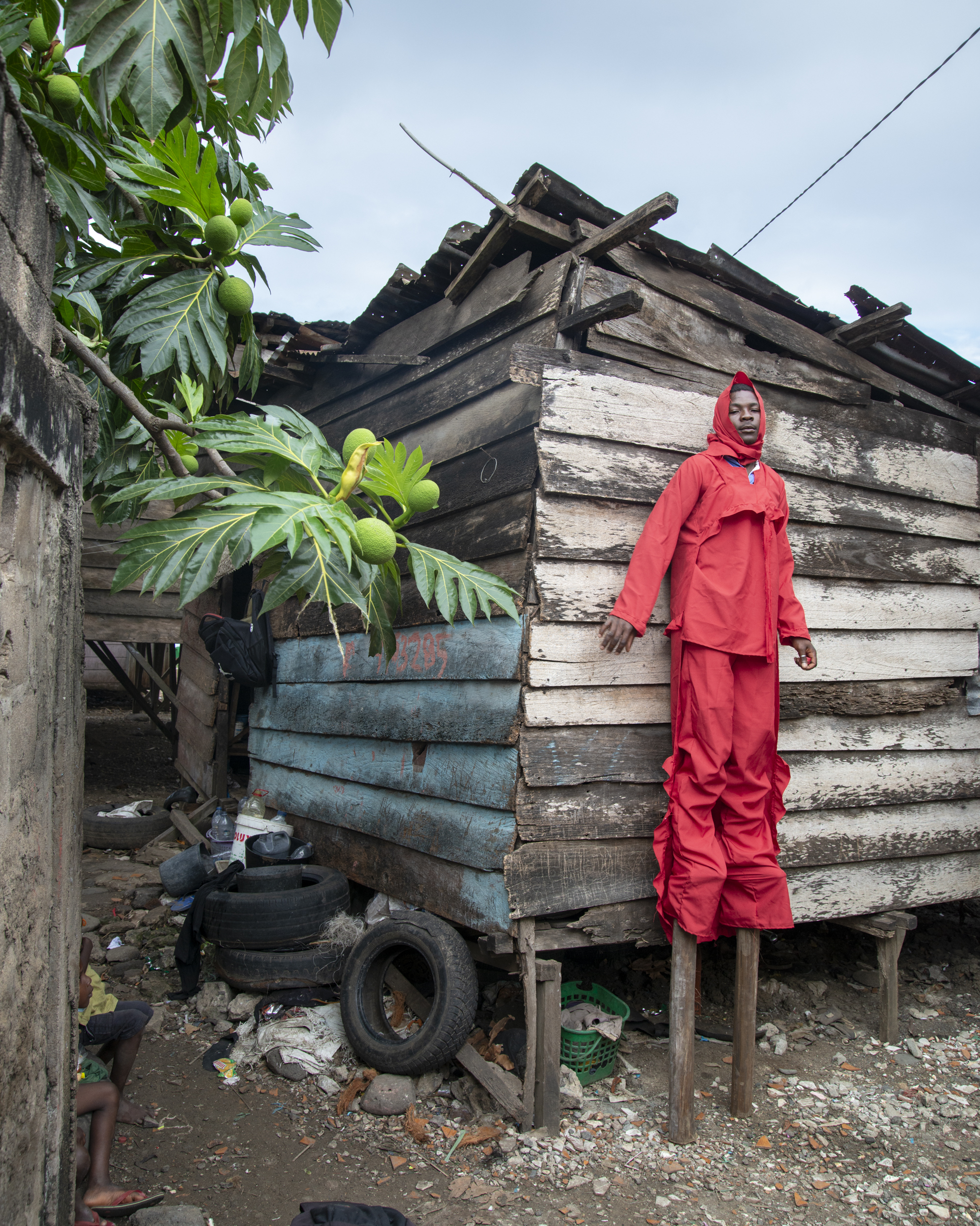 Person in bright red protective suit standing beside weathered wooden house on stilts with tropical plants and tyre visible.