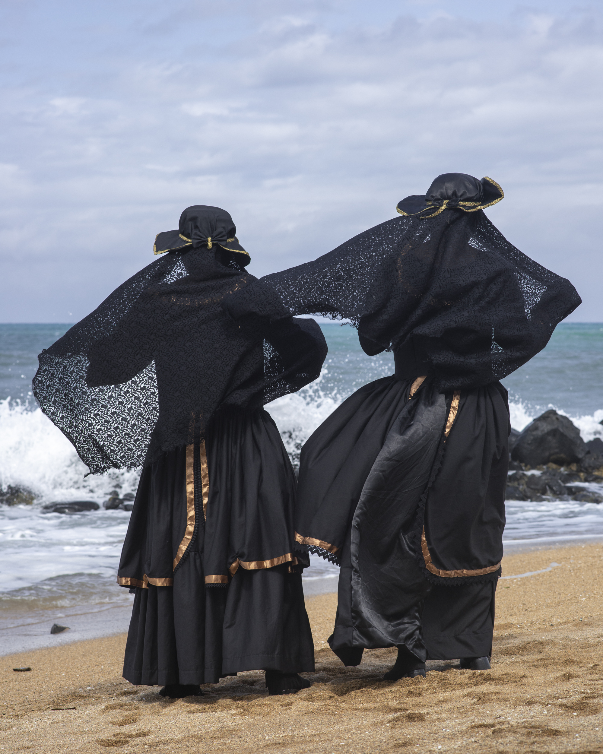 Two women in traditional black clothing with lace shawls and bonnets standing on beach, facing ocean waves under cloudy sky.
