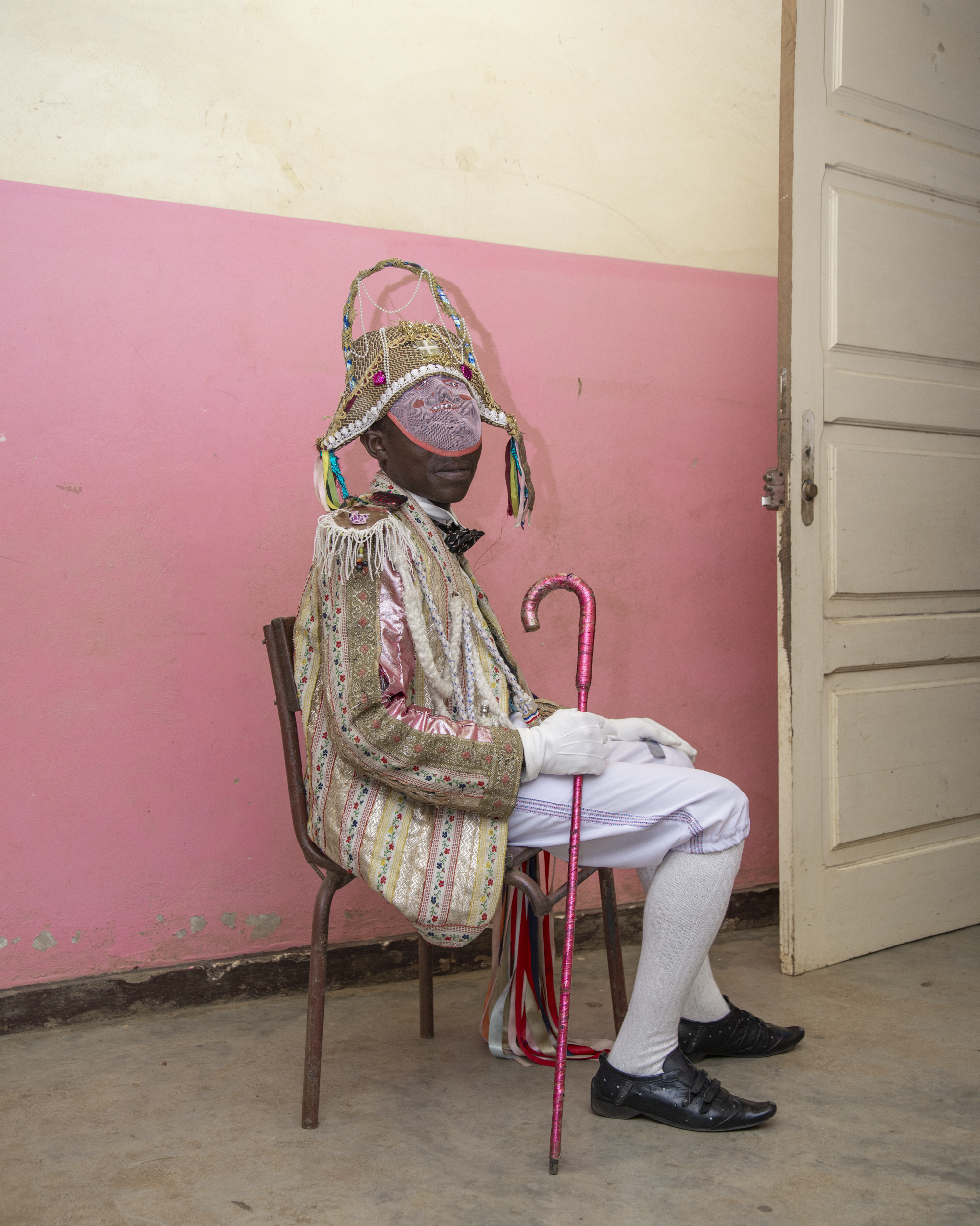 Person in ornate gold and silver garments with elaborate headdress sits on brown chair, holding red walking stick against pink and cream walls.