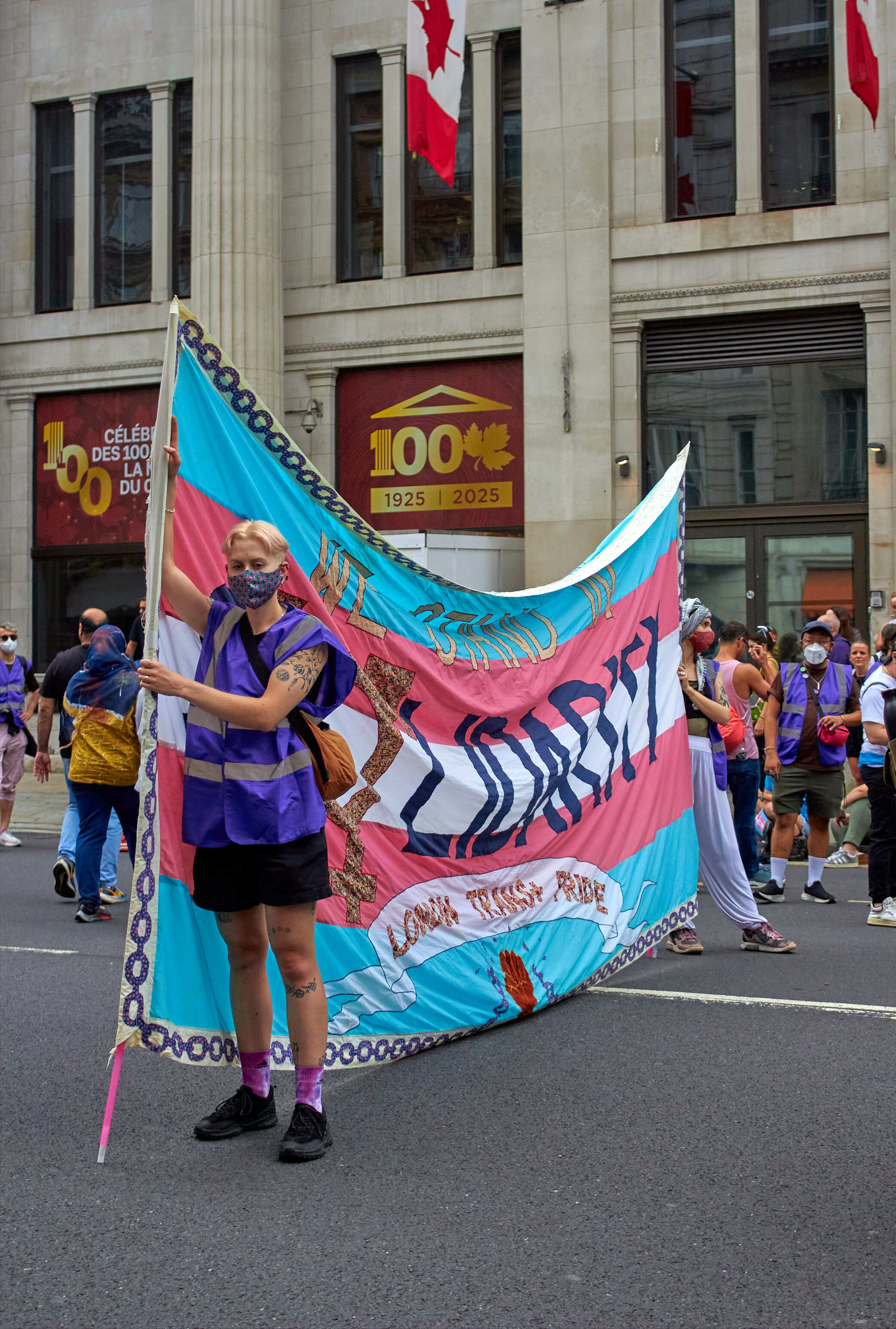 Person holding large turquoise and pink banner with black text and illustrations during street demonstration outside classical building.