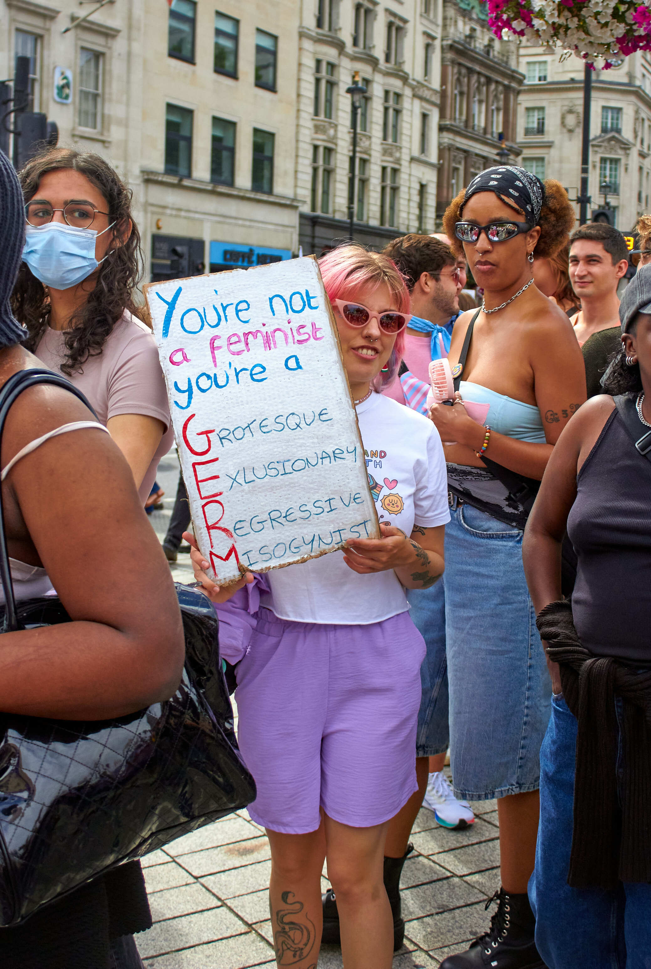 Protesters holding signs including "You're not a feminist you're a TERF" on city street with historic buildings in background.