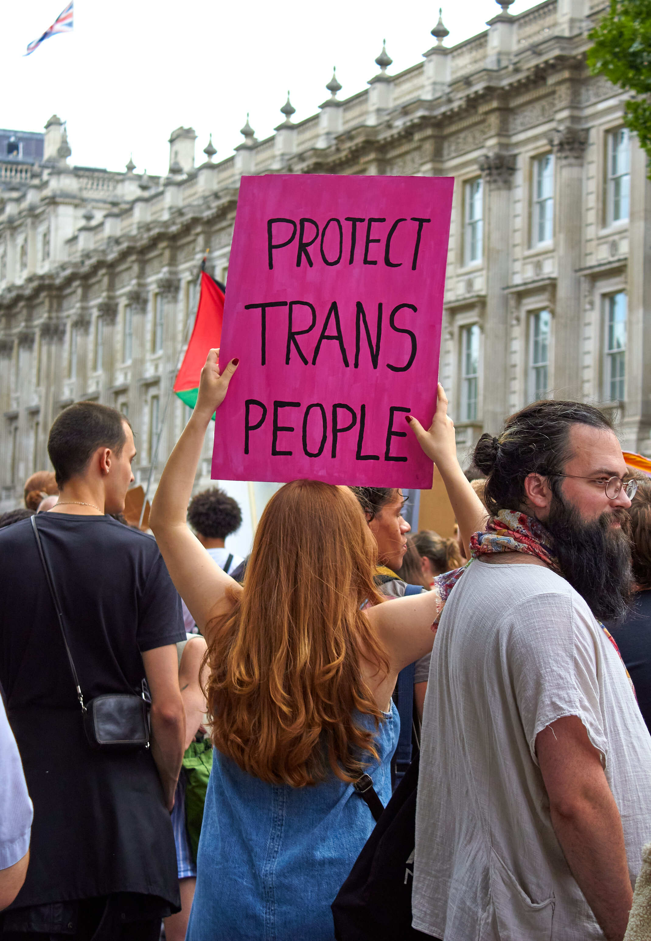 Crowd holding bright pink protest sign reading "PROTECT TRANS PEOPLE" in front of ornate stone government building with flag.