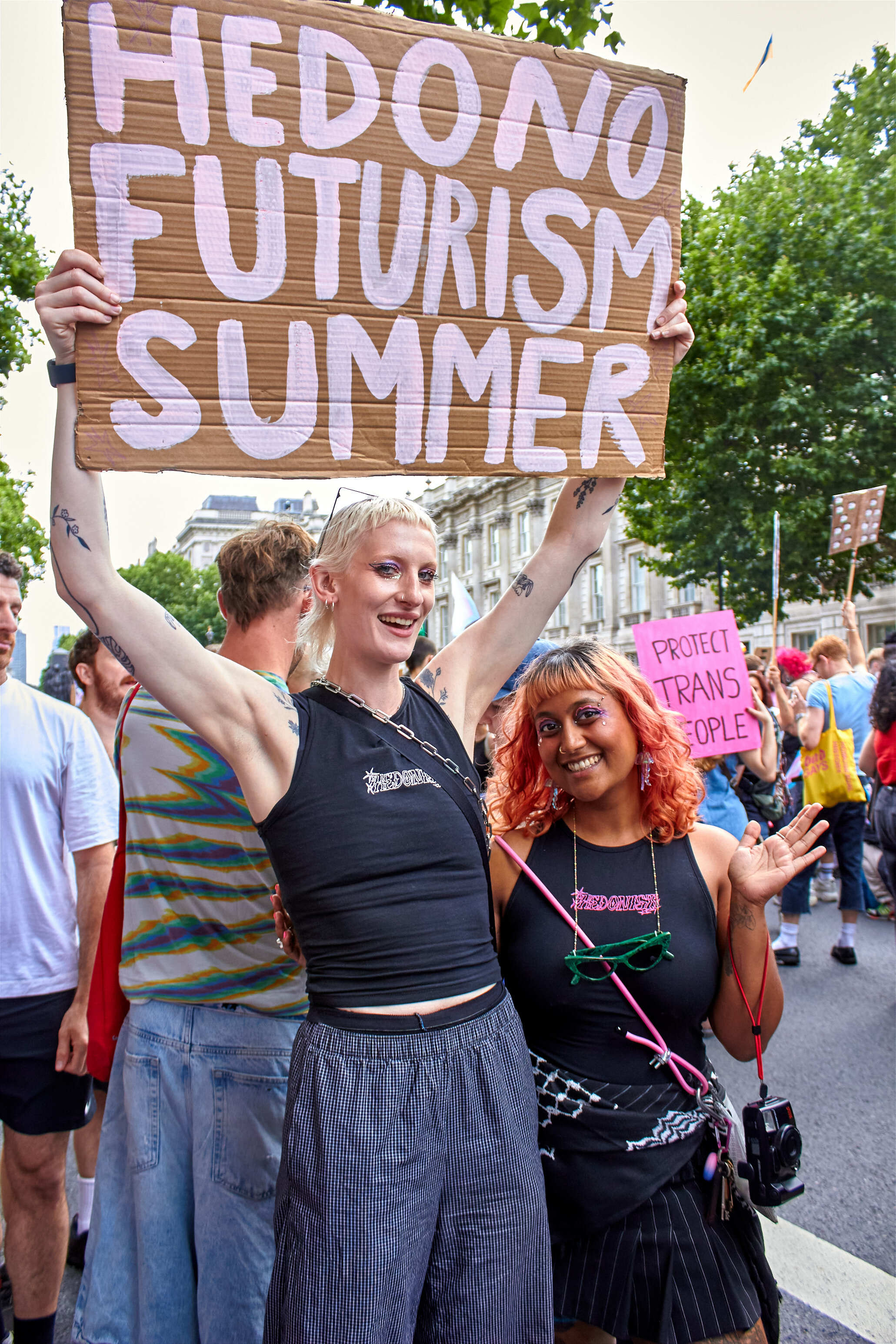 Two people at outdoor demonstration holding brown cardboard sign with white text "HEDONIC FUTURISM SUMMER", surrounded by crowd.