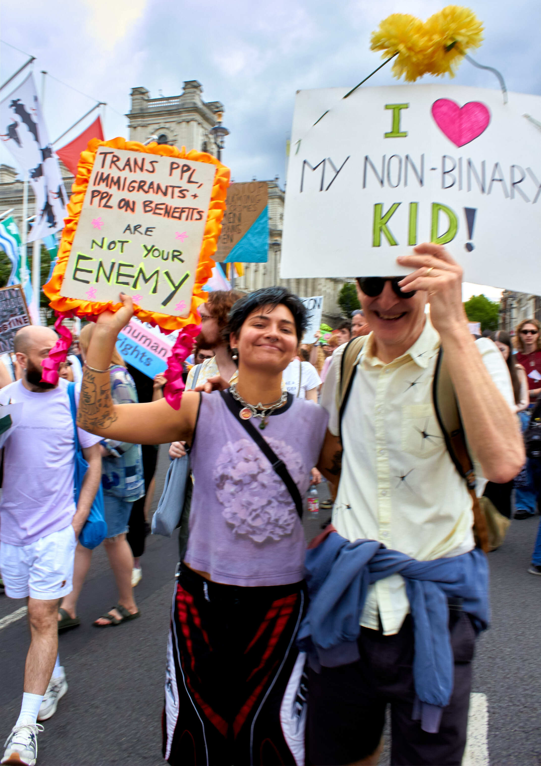 Two protesters holding signs reading "TRANS PPL, IMMIGRANTS, PPL ON BENEFITS ARE NOT YOUR ENEMY" and "I ♥ MY NON-BINARY KID!" at outdoor demonstration.