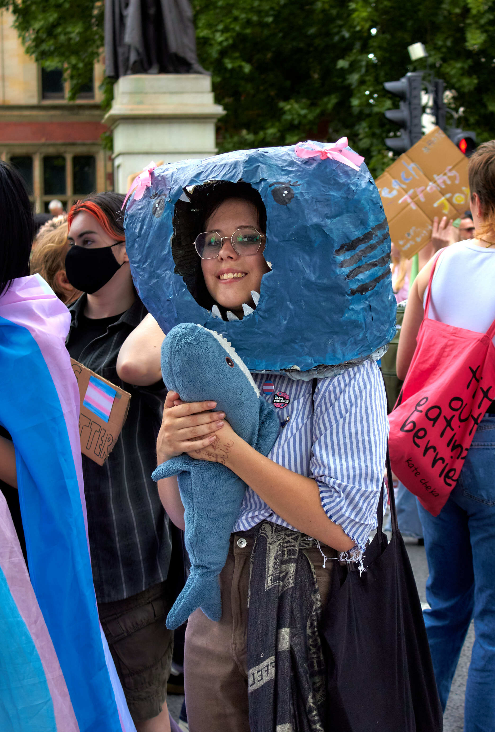 Person at outdoor protest wearing large blue shark costume head with open mouth showing teeth, holding matching blue tail.