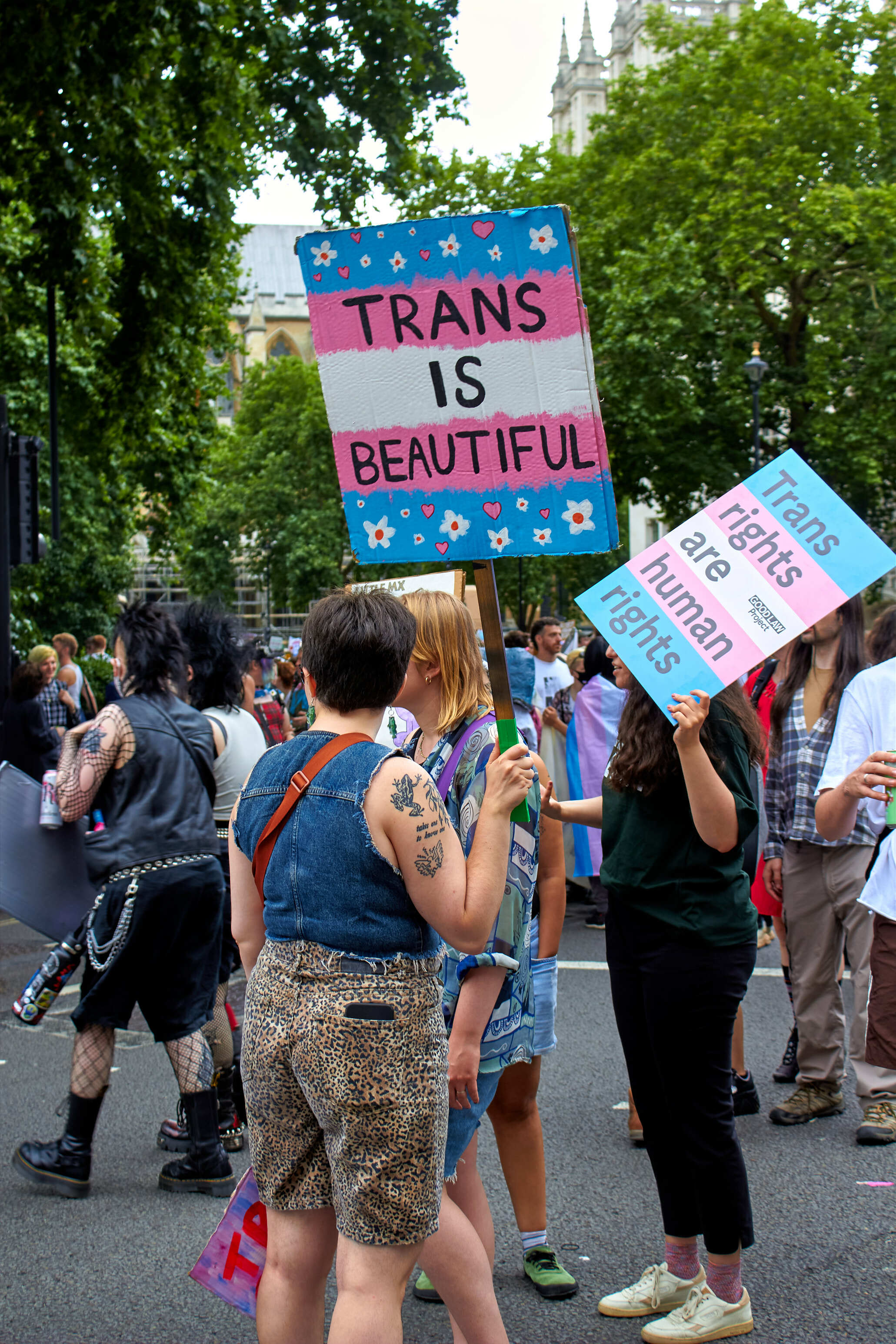 Protesters holding colourful signs reading "TRANS IS BEAUTIFUL" in blue, pink and white colours with floral decorations at outdoor demonstration.
