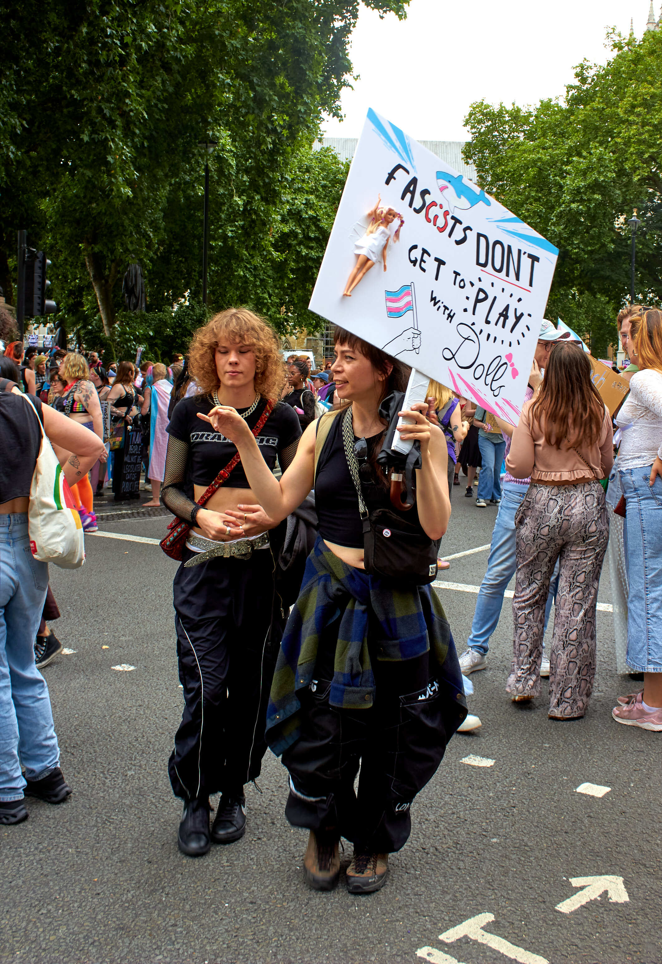 Two people at street demonstration holding white placard with colourful text about fascists, surrounded by crowd on tarmac road.