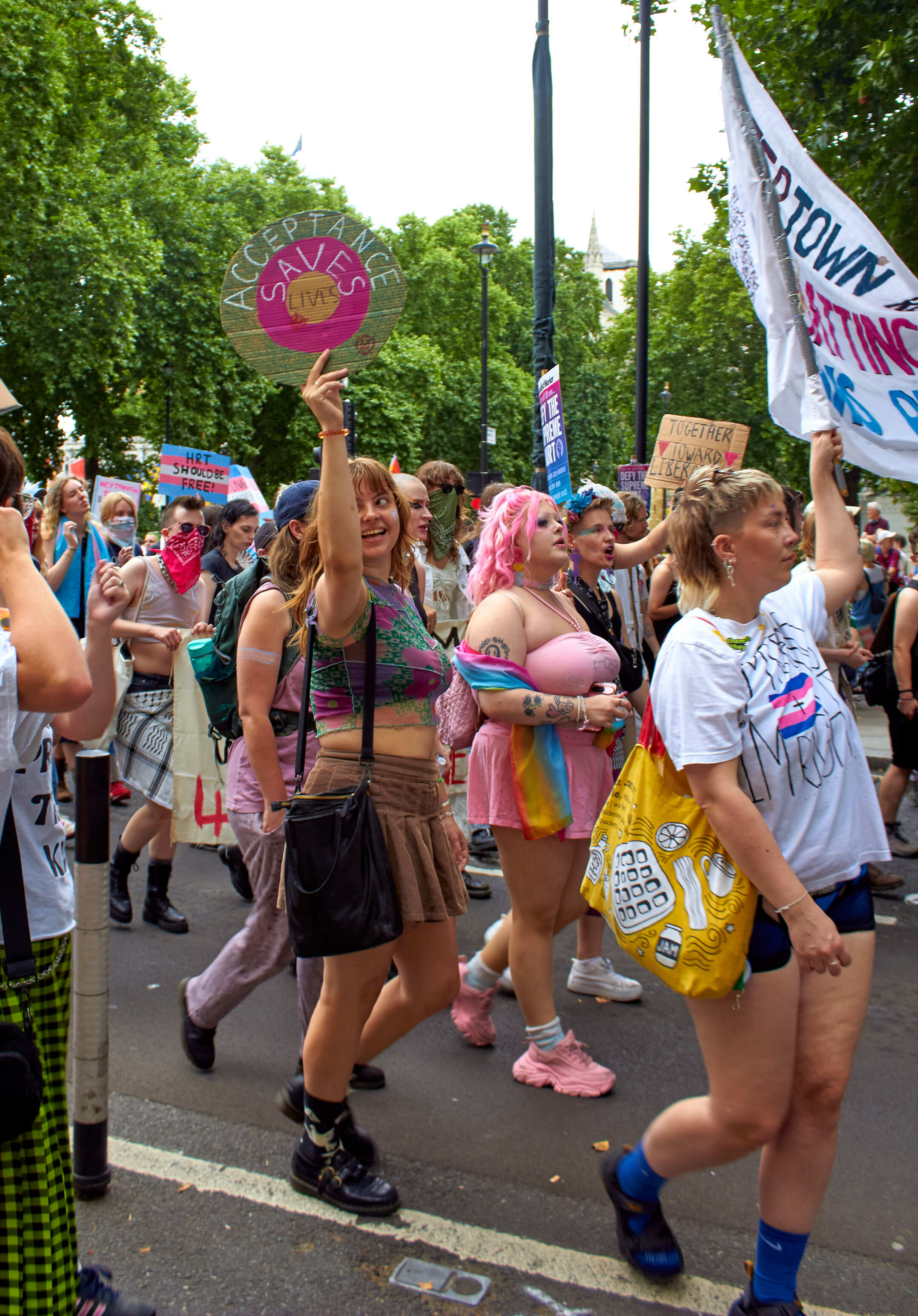 Crowd of protesters on tree-lined street holding placards and banners, wearing colourful clothing and pink accessories.