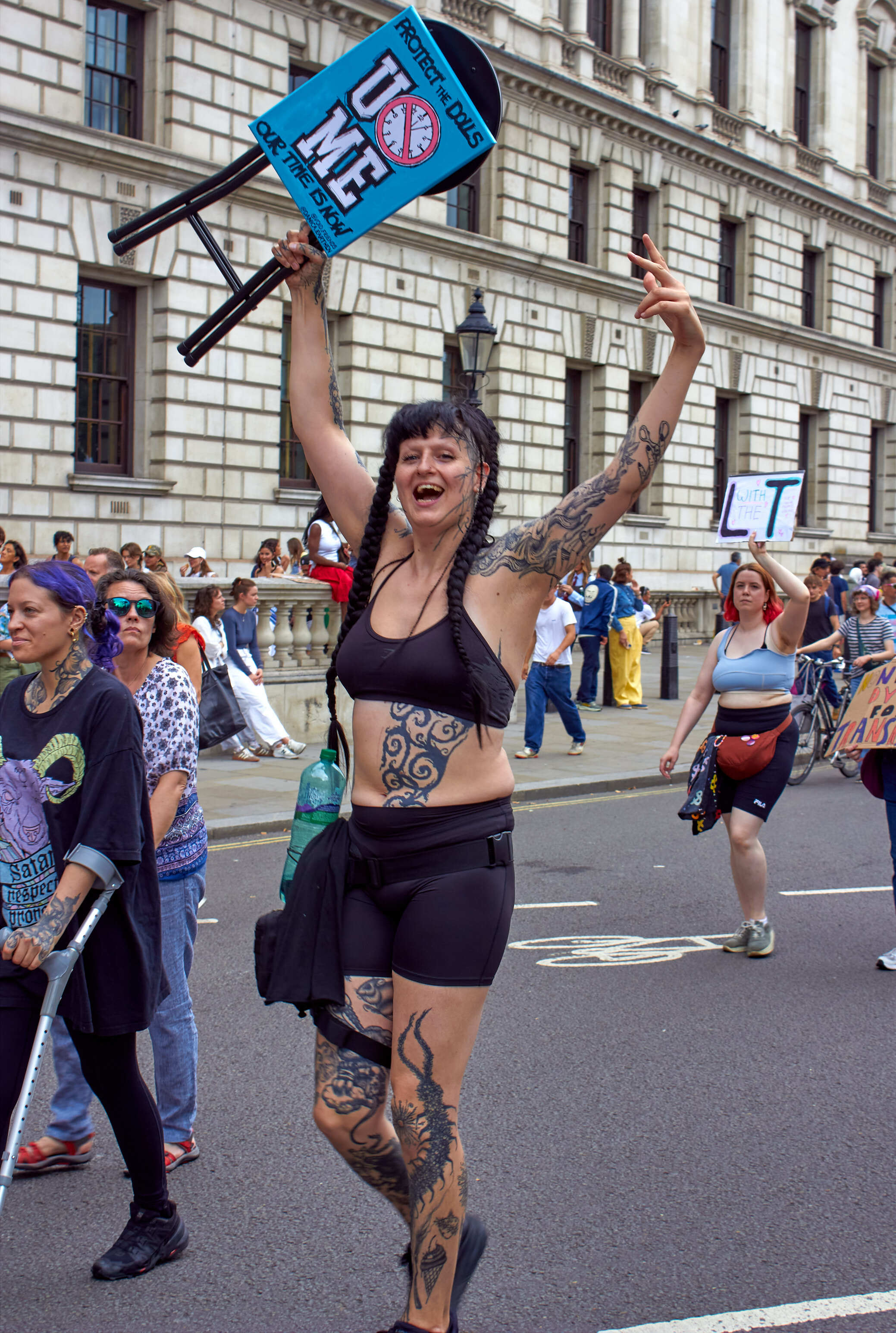 Woman in black bikini with tattoos holds blue "SAVE THE NHS" banner above head during street protest outside stone government building.
