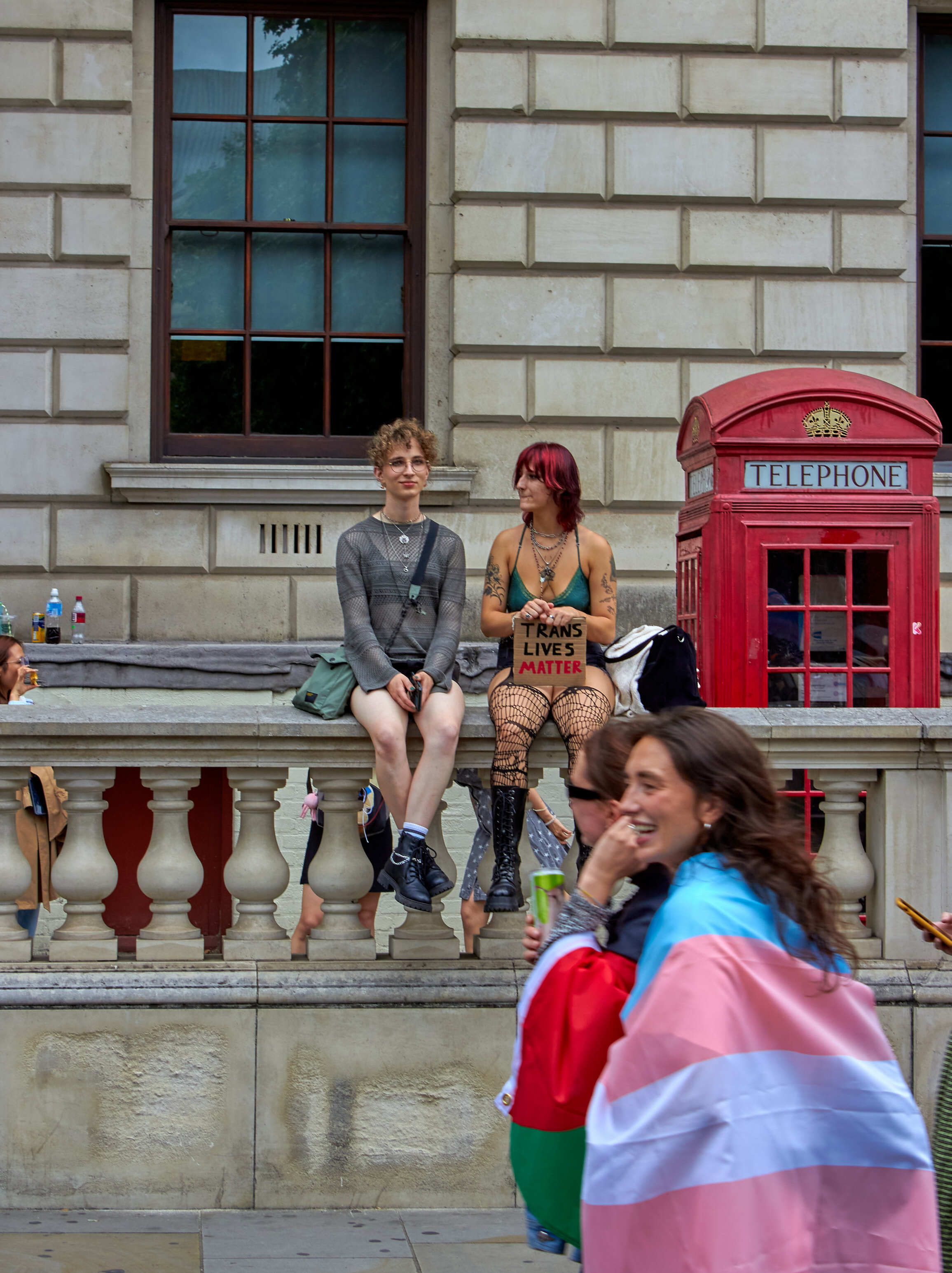 Three people on stone steps by red telephone box and classical building. Person in foreground wrapped in pink and blue striped flag.