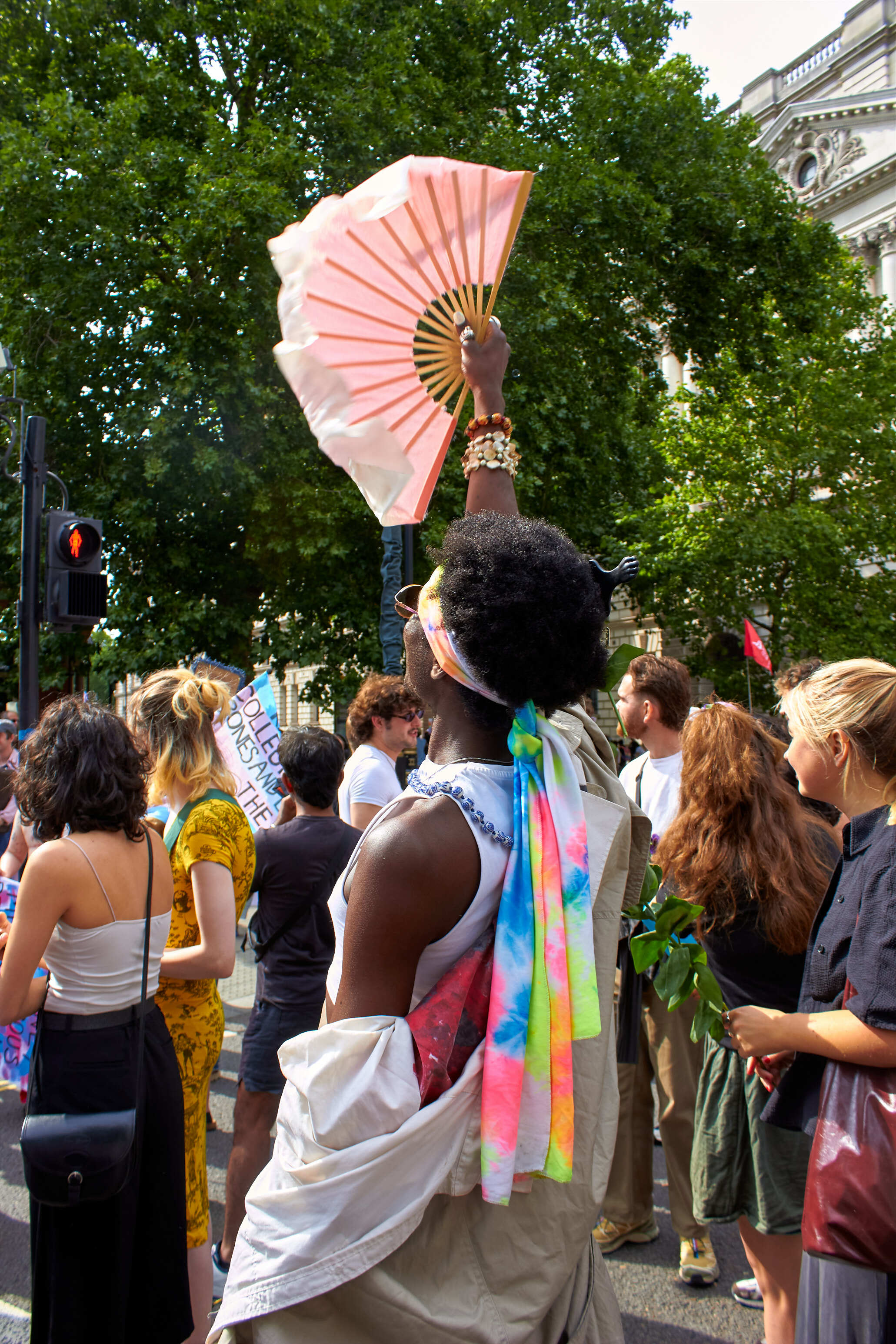 Person holding pink fan aloft in crowd, wearing rainbow ribbons and white wrap, surrounded by protesters near leafy trees and building.