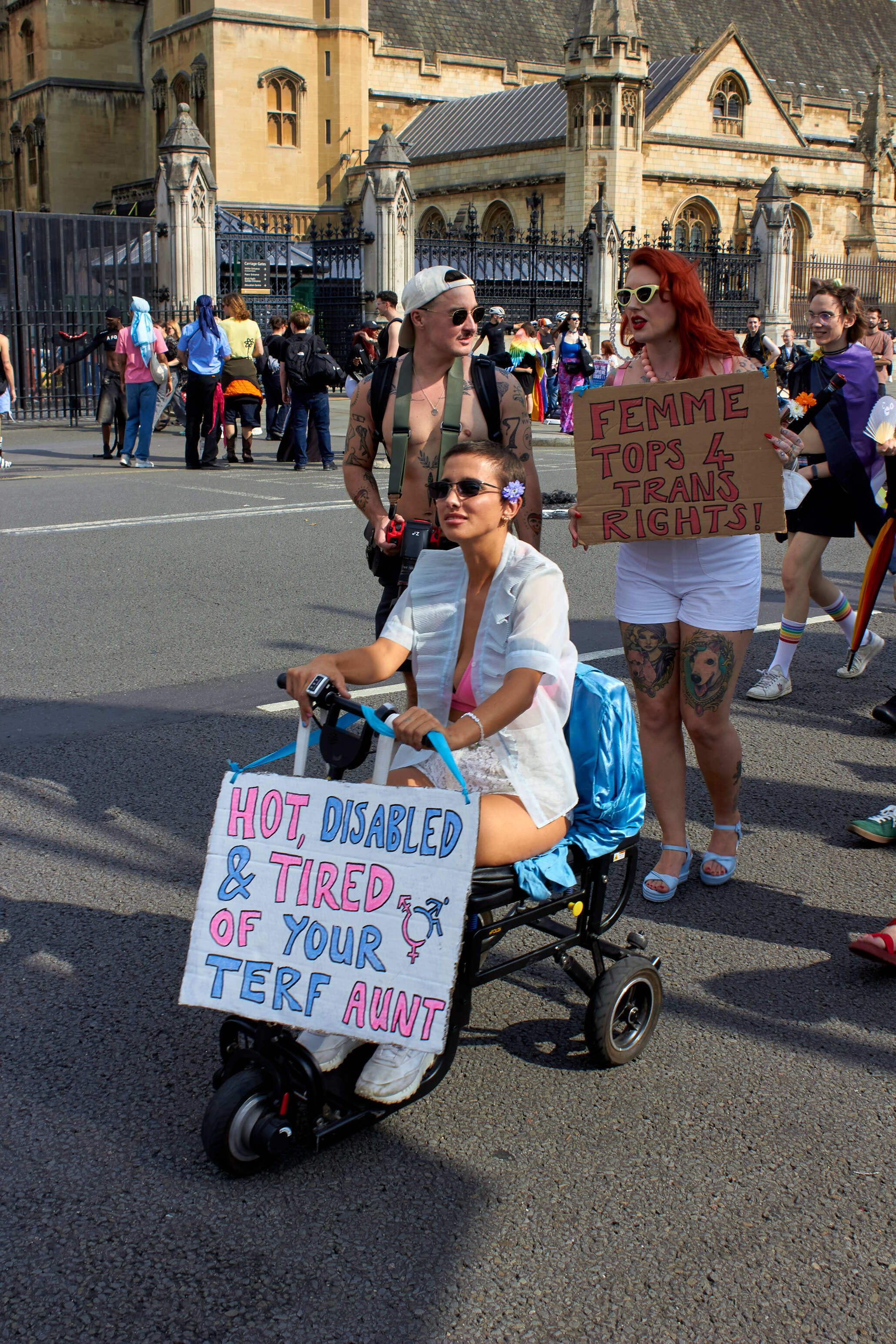 Protesters on street with placards, woman in wheelchair holding sign reading "HOT DISABLED & TIRED OF YOUR TERF ANNT", stone buildings behind.