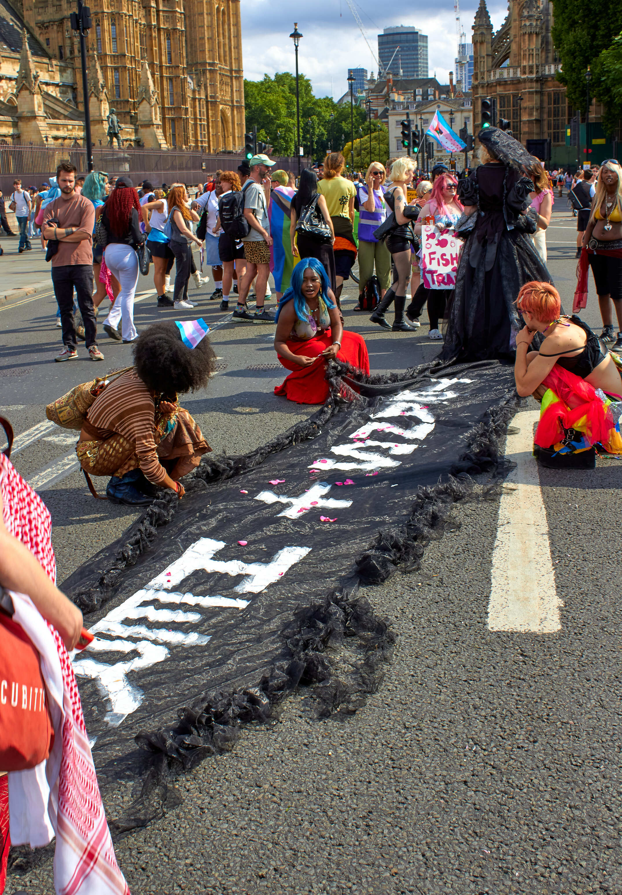 Street protest with large black banner displaying white text, surrounded by demonstrators near Westminster buildings and modern skyscrapers.
