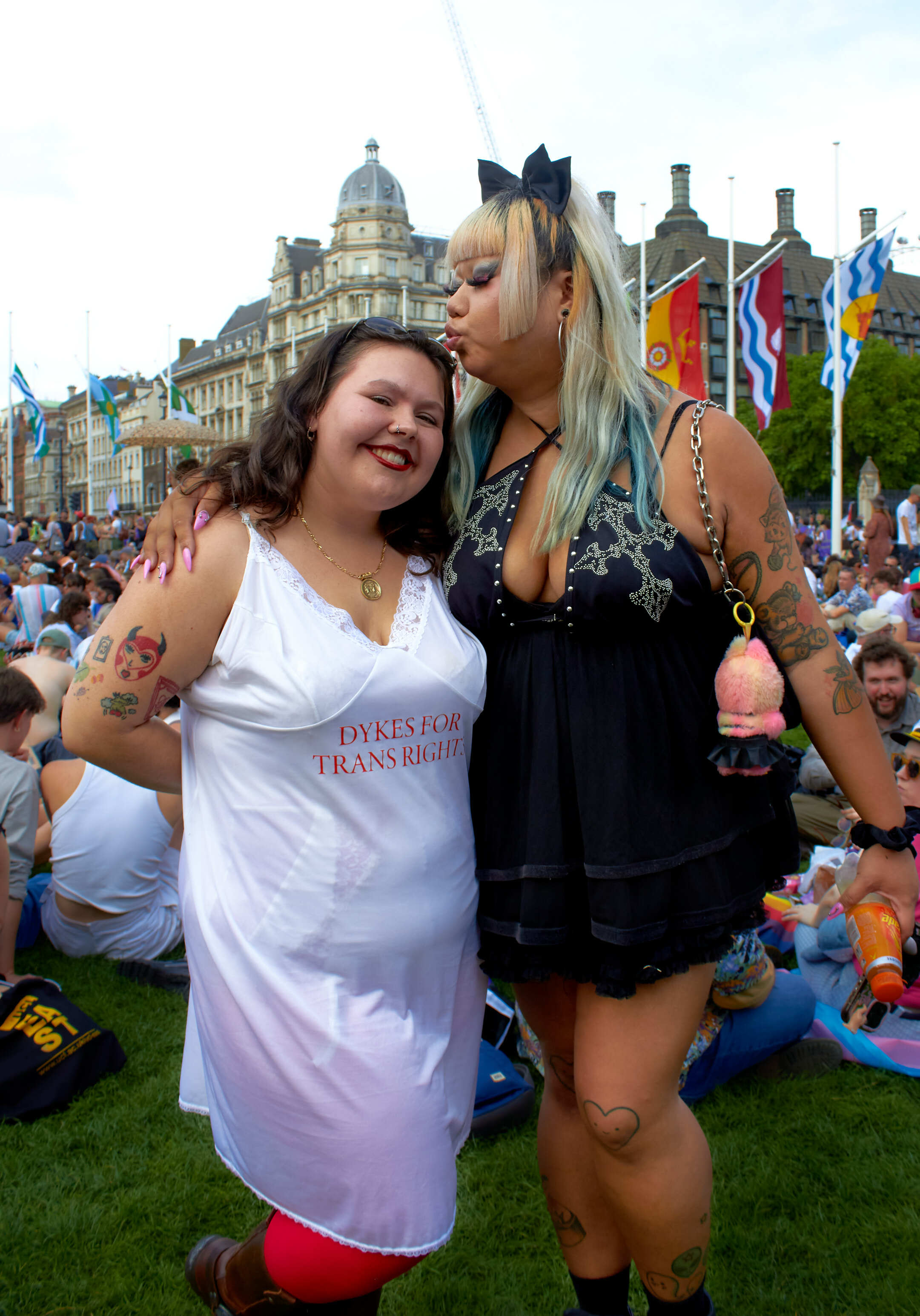 Two women embracing at outdoor Pride event with rainbow flags, historic buildings, and crowds in background on grass.