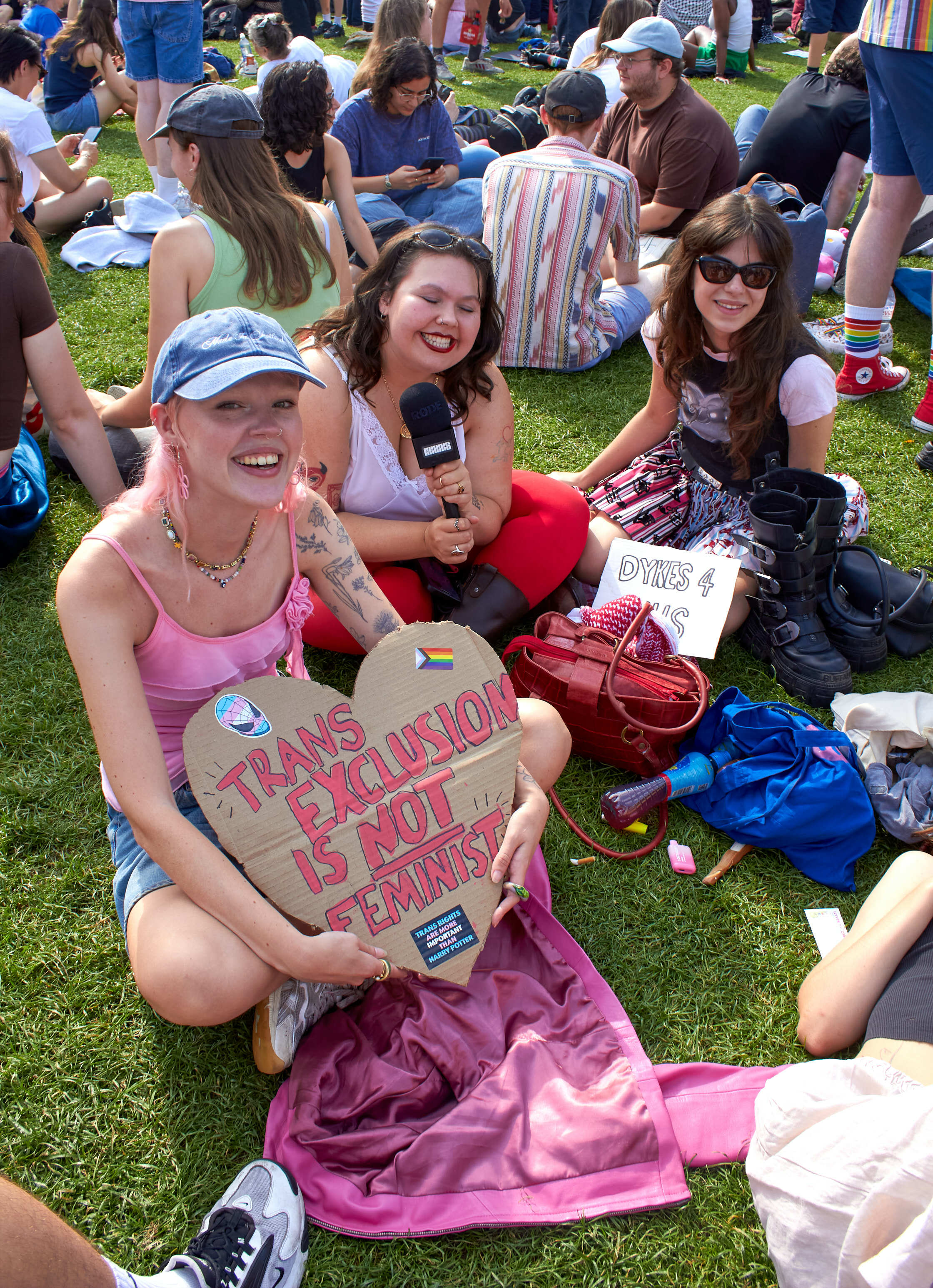 Three women sitting on grass at outdoor event, one holding heart-shaped sign reading "Trans exclusion is not feminism"