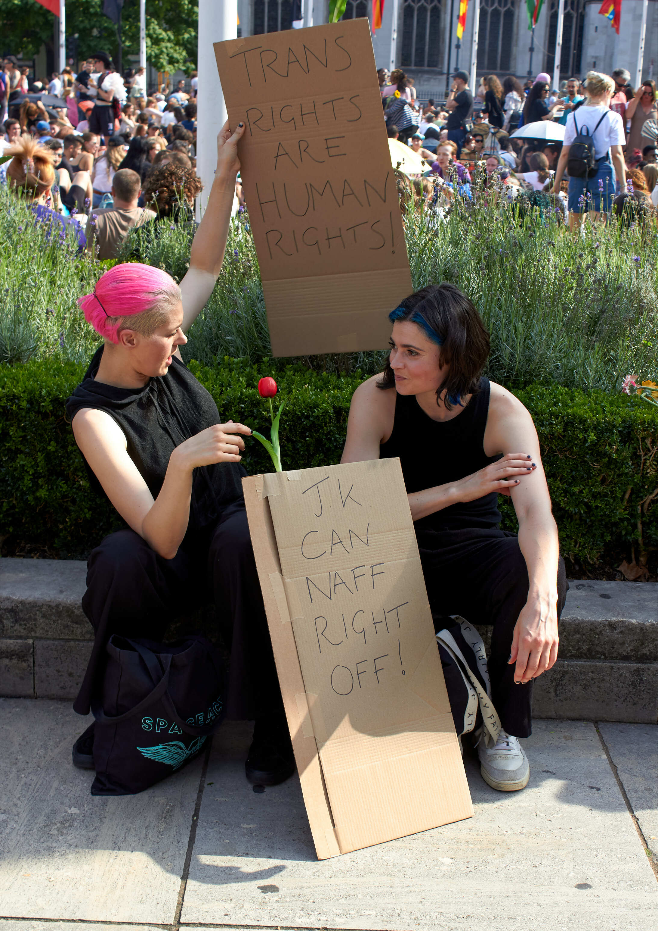 Two women in black clothing sit on steps holding cardboard protest signs, with person in pink hat holding red flower. Crowd visible behind.