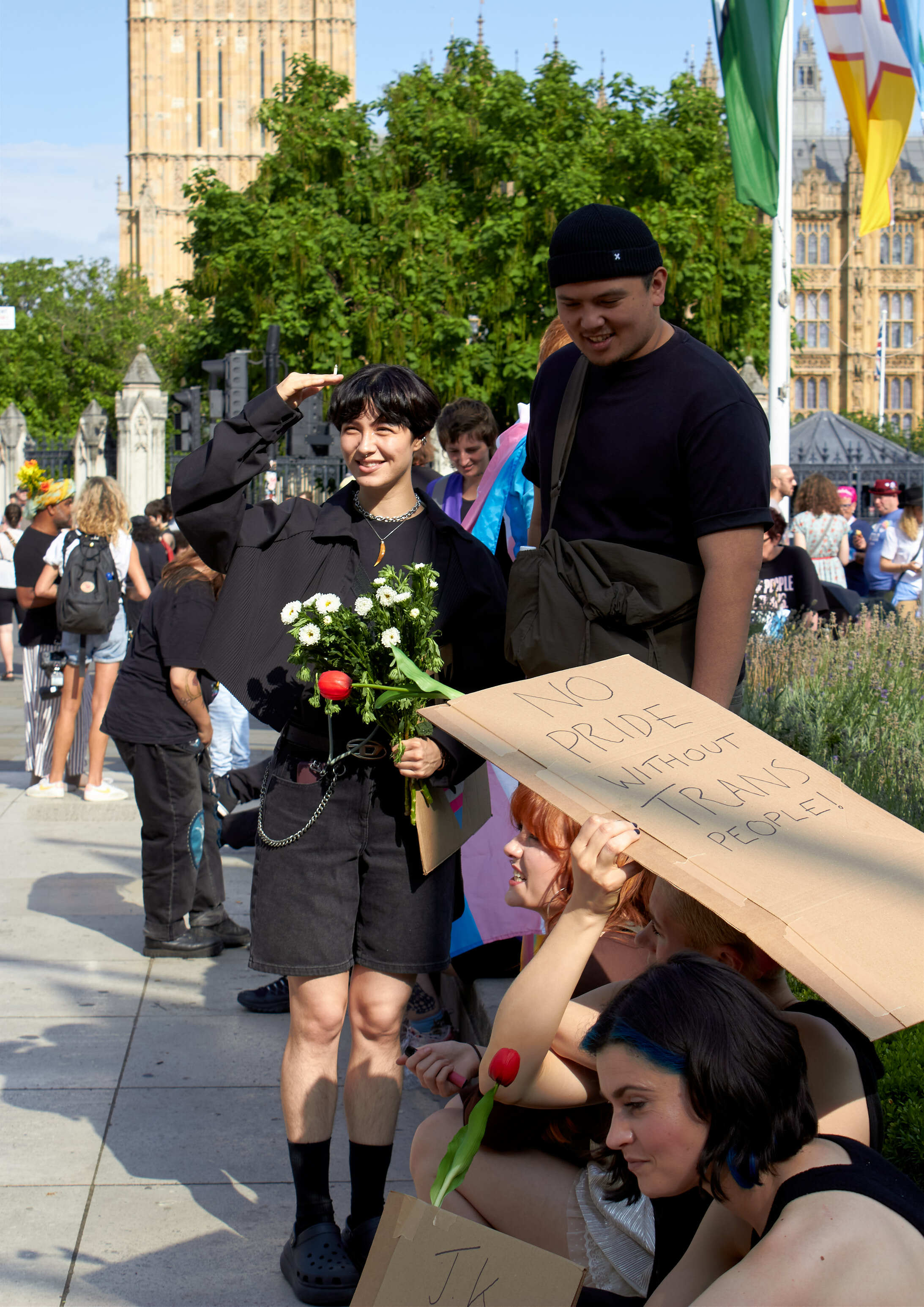 Woman in black holding white flowers stands with man in black t-shirt and beanie, with protesters holding cardboard signs nearby.