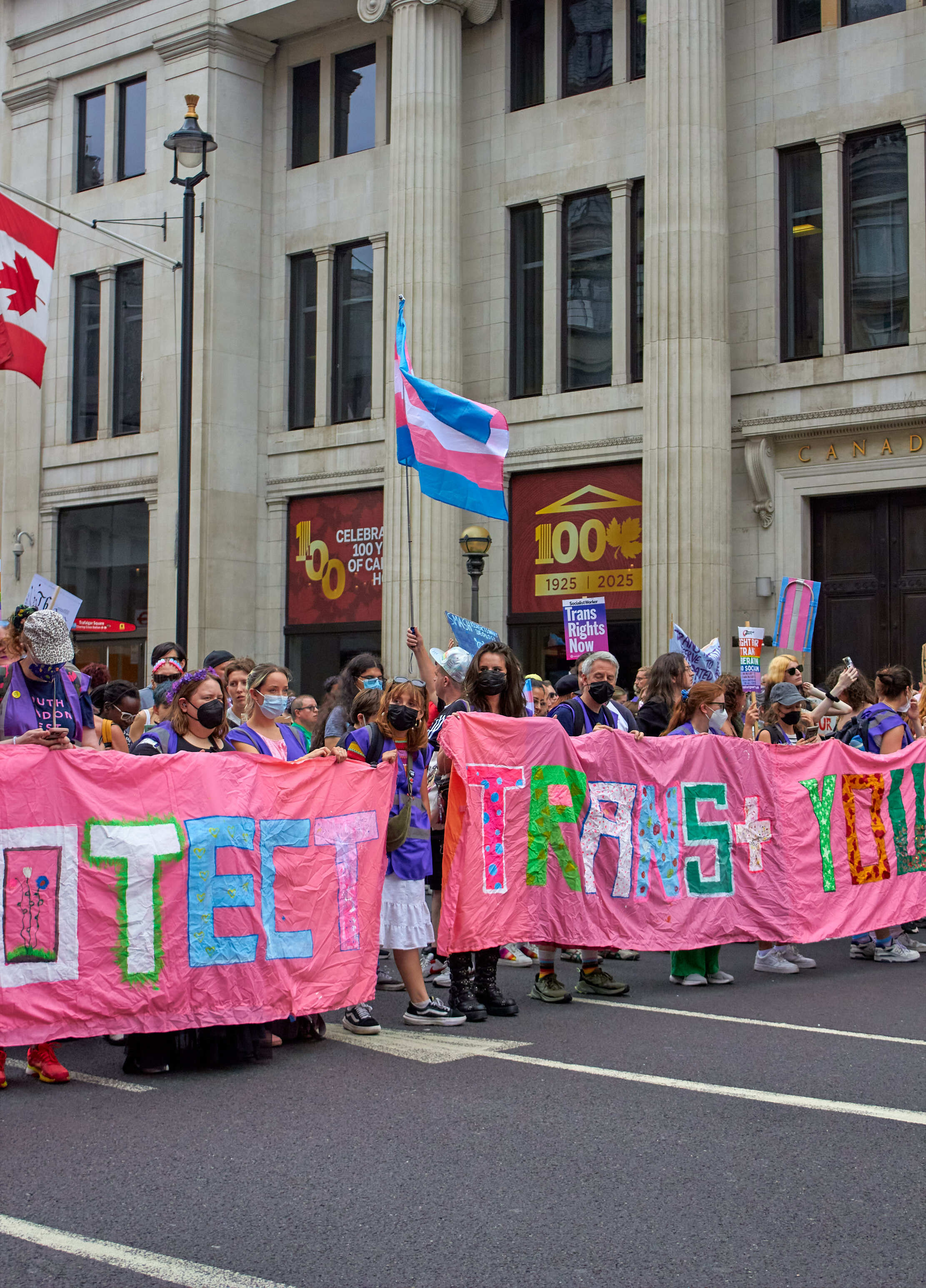 Crowd holding large pink banner with colourful letters spelling "TRANS RIGHTS" outside grey stone building with columns and flags.