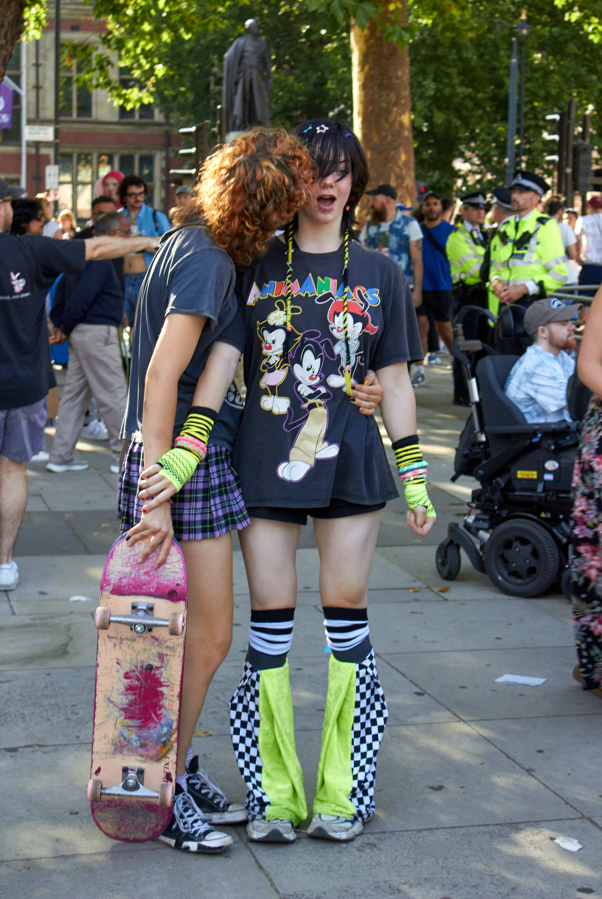 Two young people in colourful alternative clothing with skateboard at outdoor gathering, police and crowd in background.