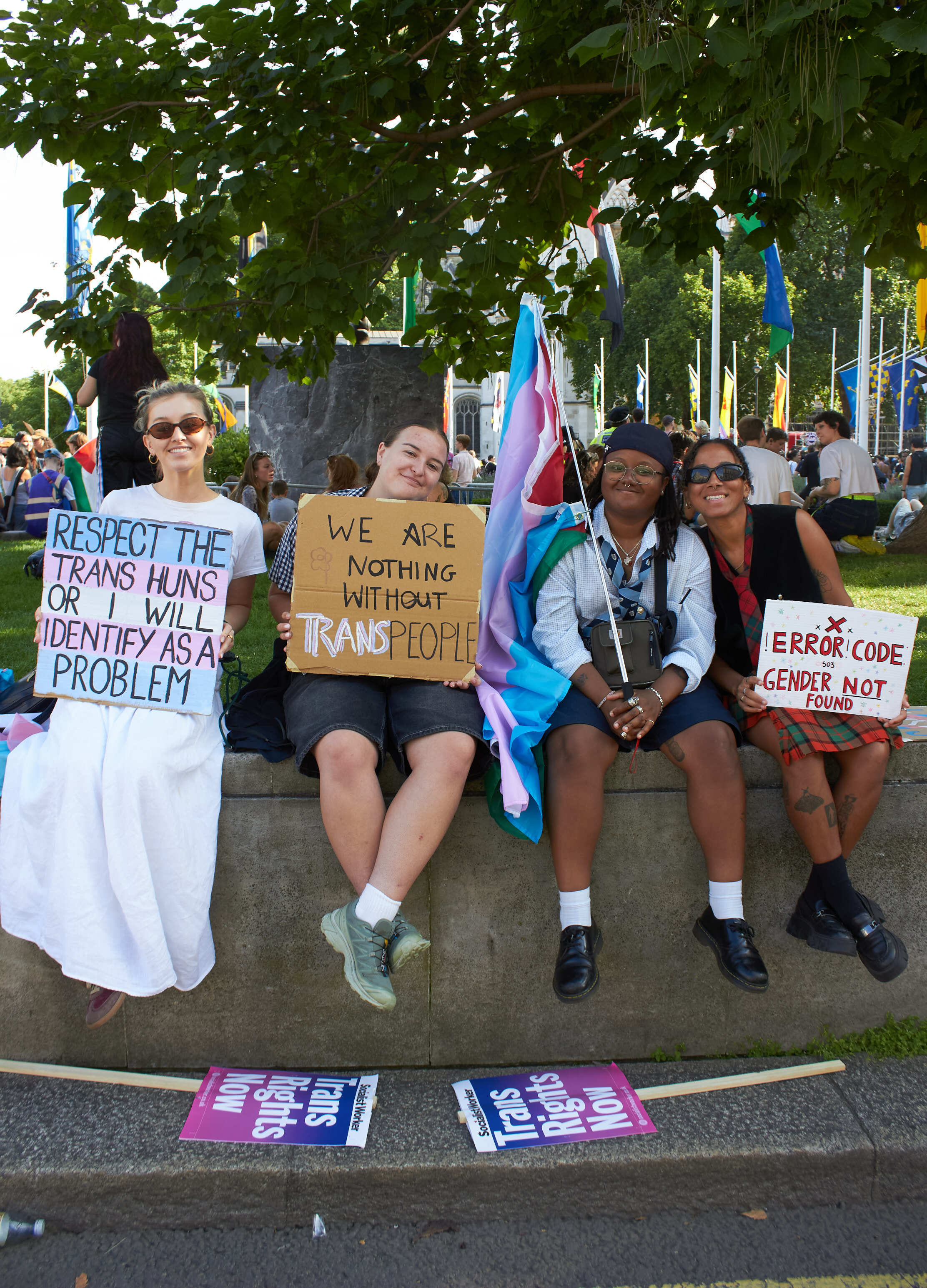 Four people sitting on concrete ledge holding protest signs supporting trans rights, with transgender flag and leafy trees overhead.