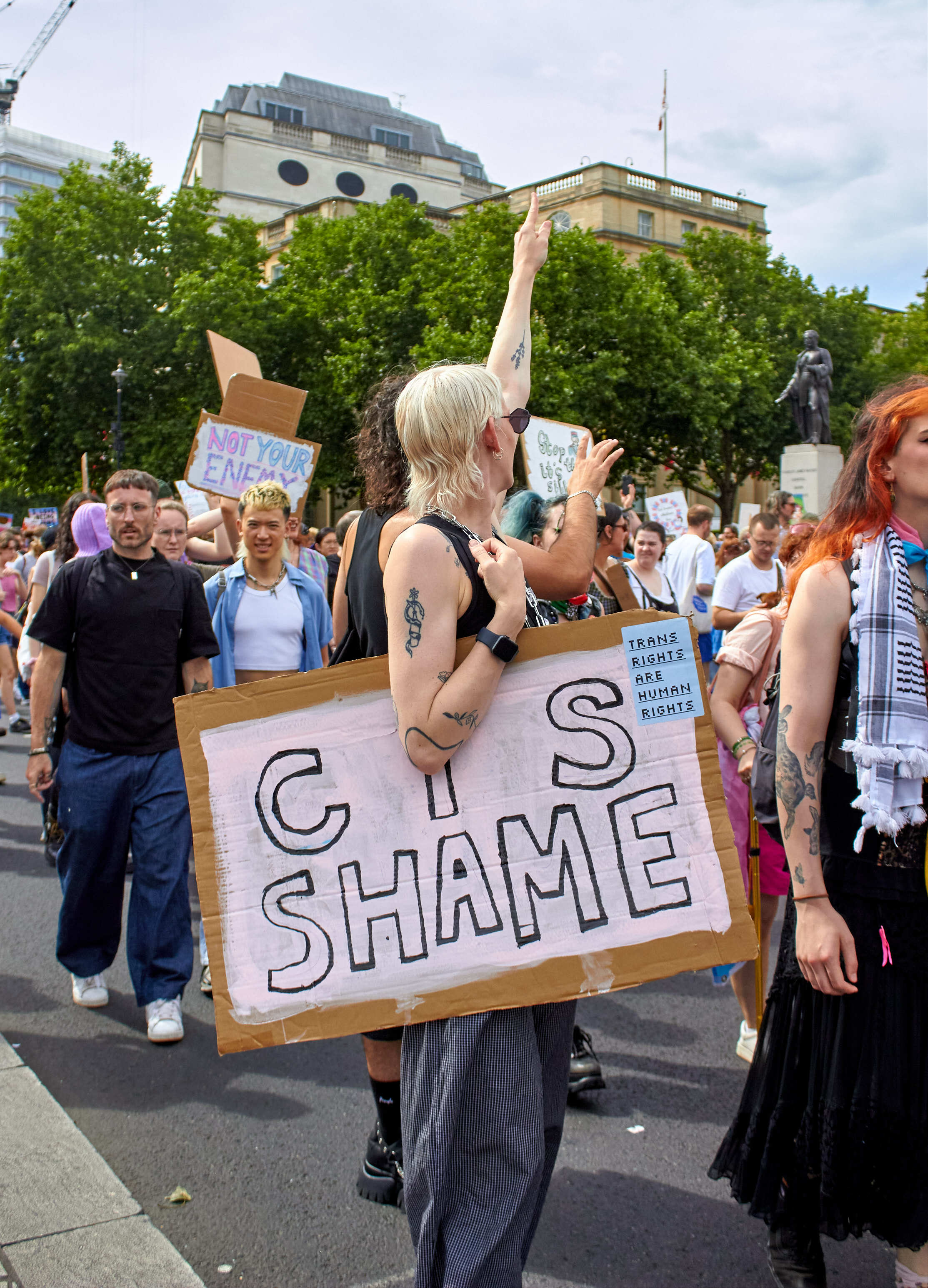 Blonde woman holding white sign reading "CIS SHAME" at outdoor protest, surrounded by crowd with placards and green trees behind.