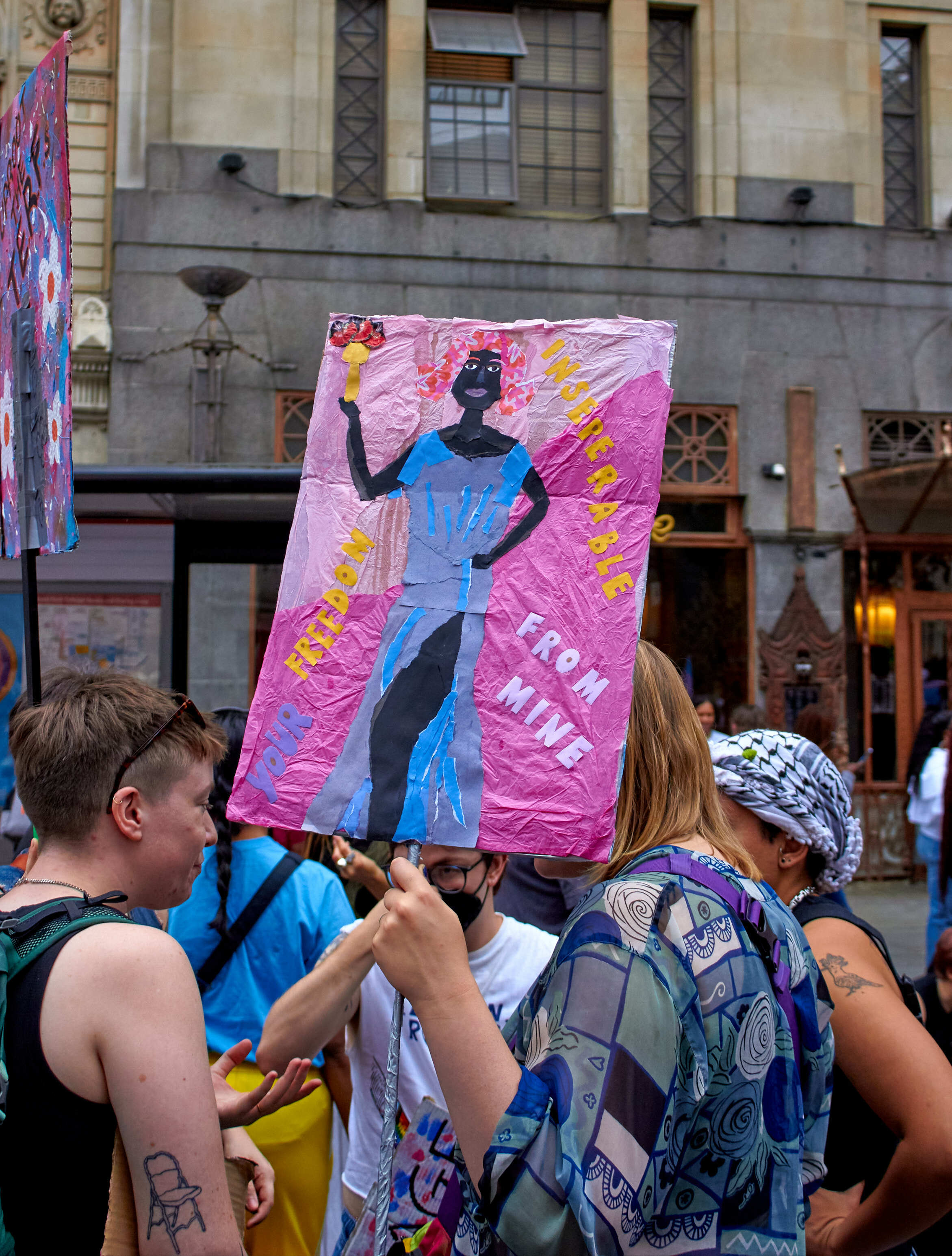 Protesters holding pink banner with blue silhouette figure and yellow text, gathered outside stone building with classical columns.