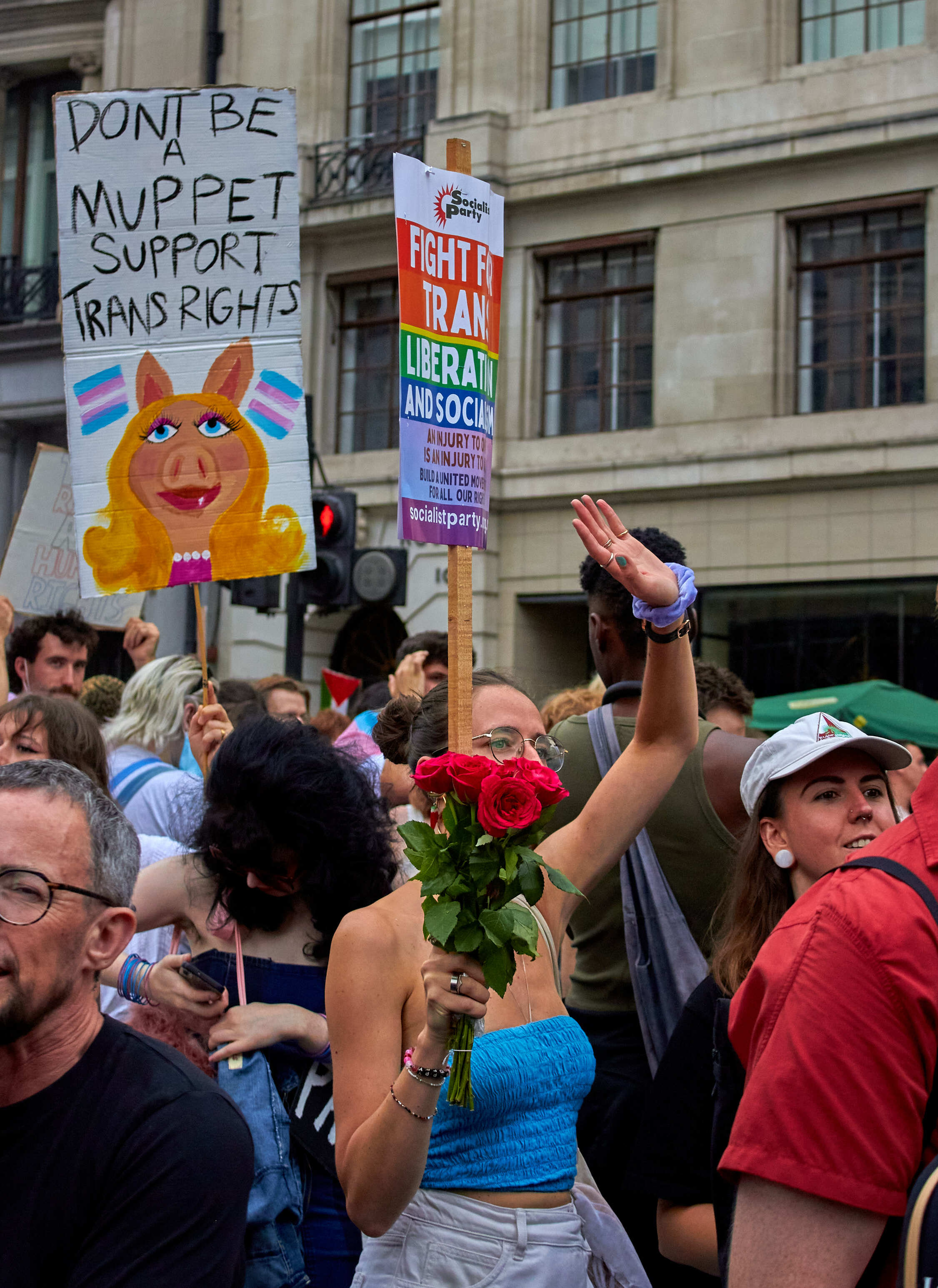 Crowd holding protest signs including one with Muppet character and "Don't be Muppet Support Trans Rights" text, person with red roses visible.