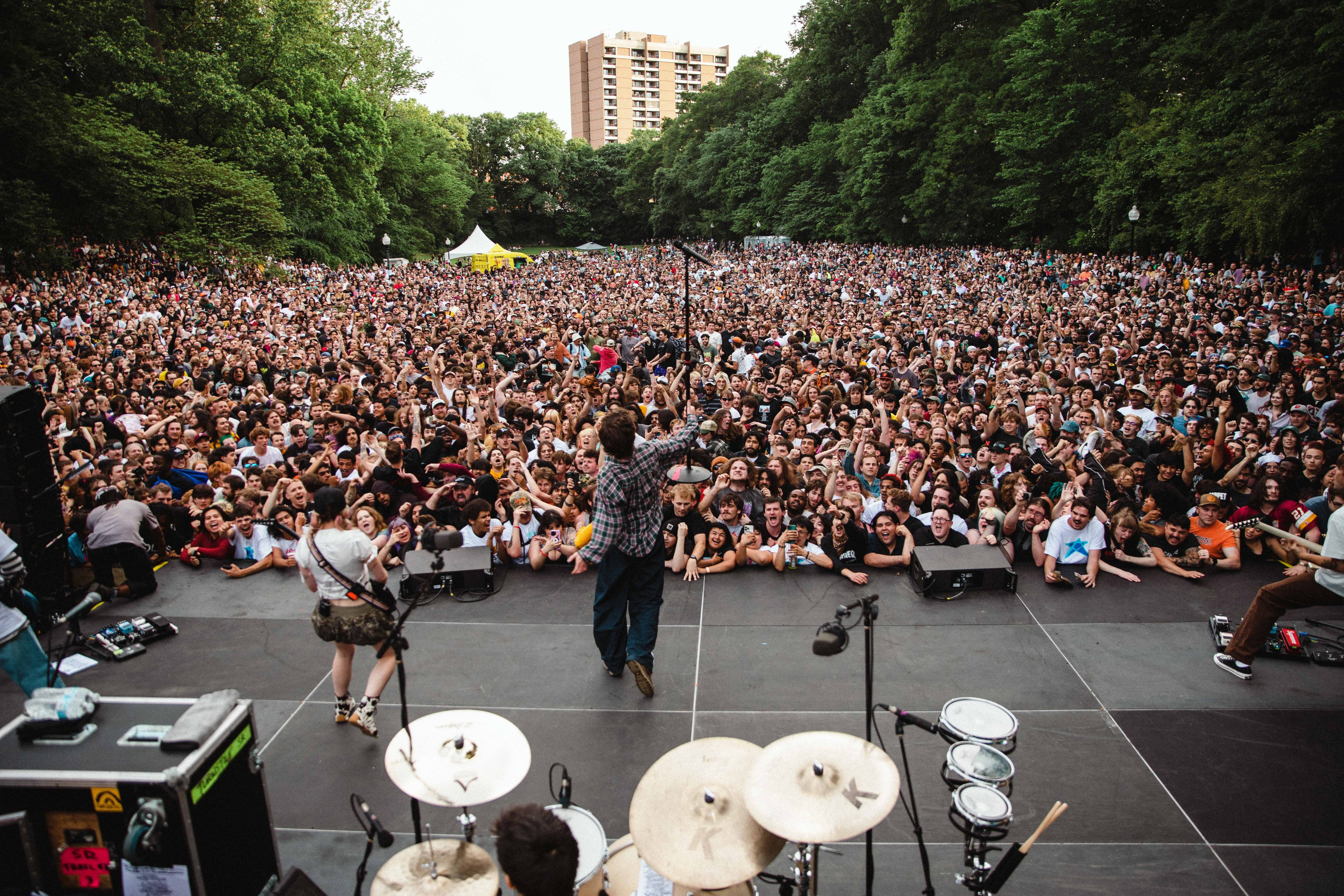 Vast crowd of people attending an outdoor music event, with a performer on stage surrounded by a sea of spectators.