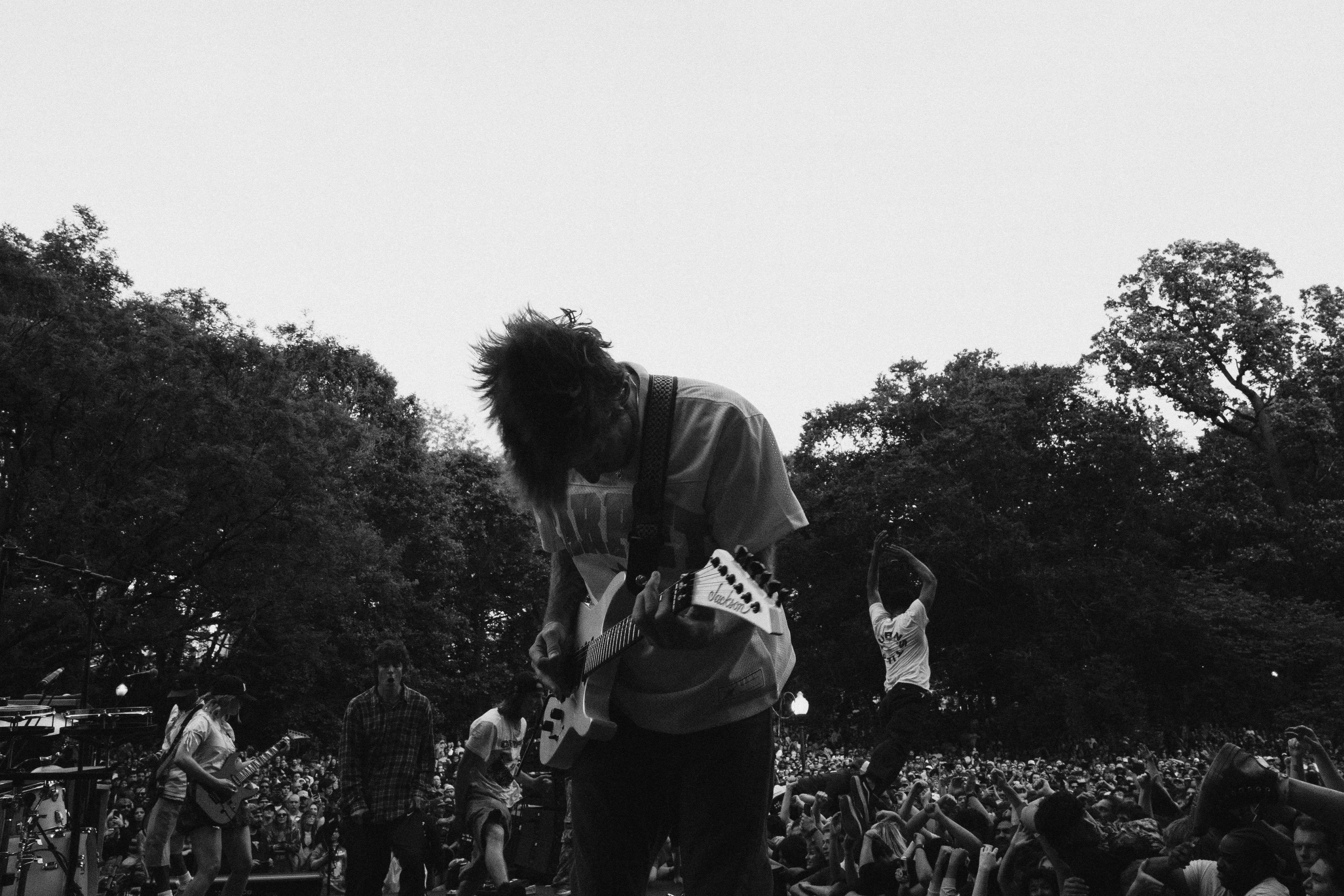 Crowd of people at an outdoor event, with a person in the centre performing on stage amidst greenery and trees.