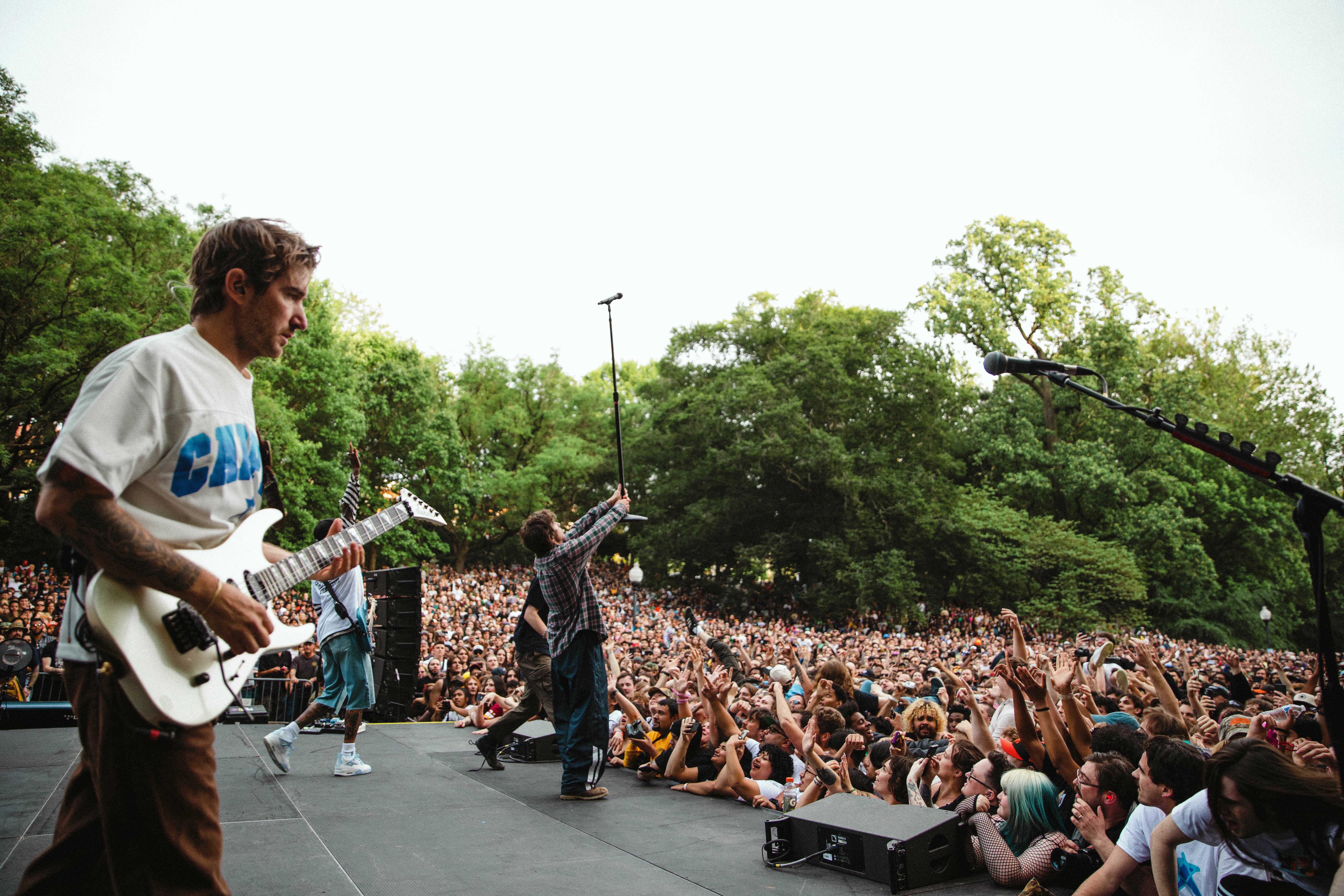 A large outdoor crowd at a music festival, with a band performing on stage amongst the trees.