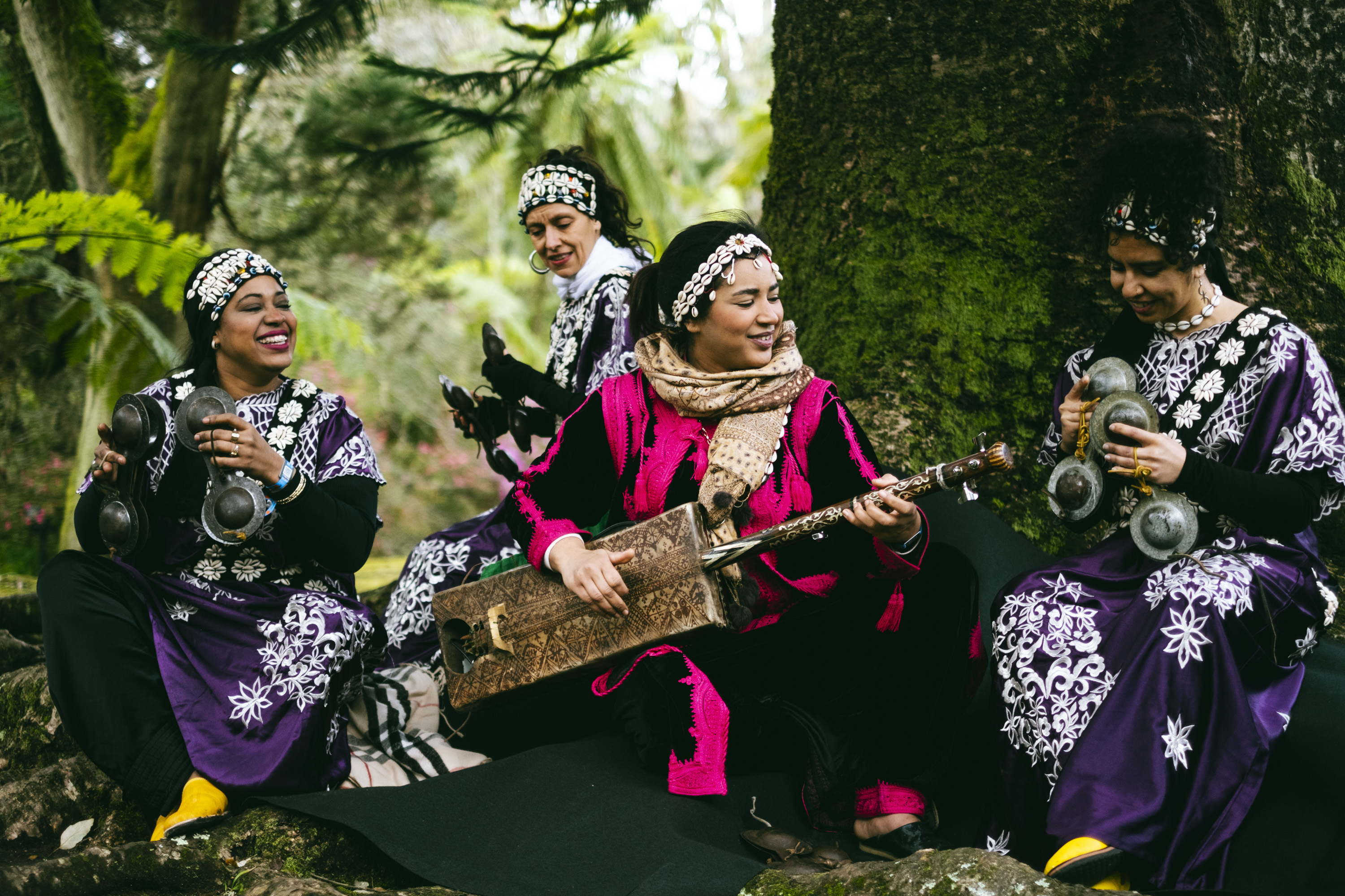 Four people wearing traditional patterned clothing, sitting in a garden setting with lush green foliage.