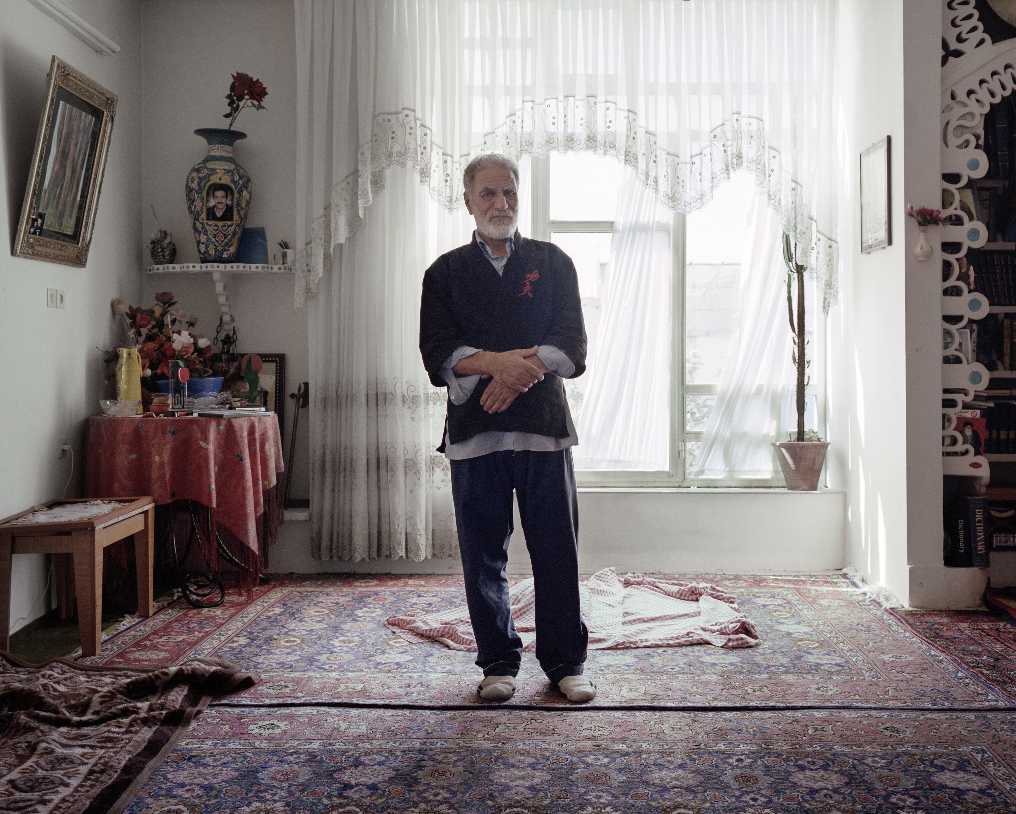 An older man standing in a room with ornate curtain and rug.
