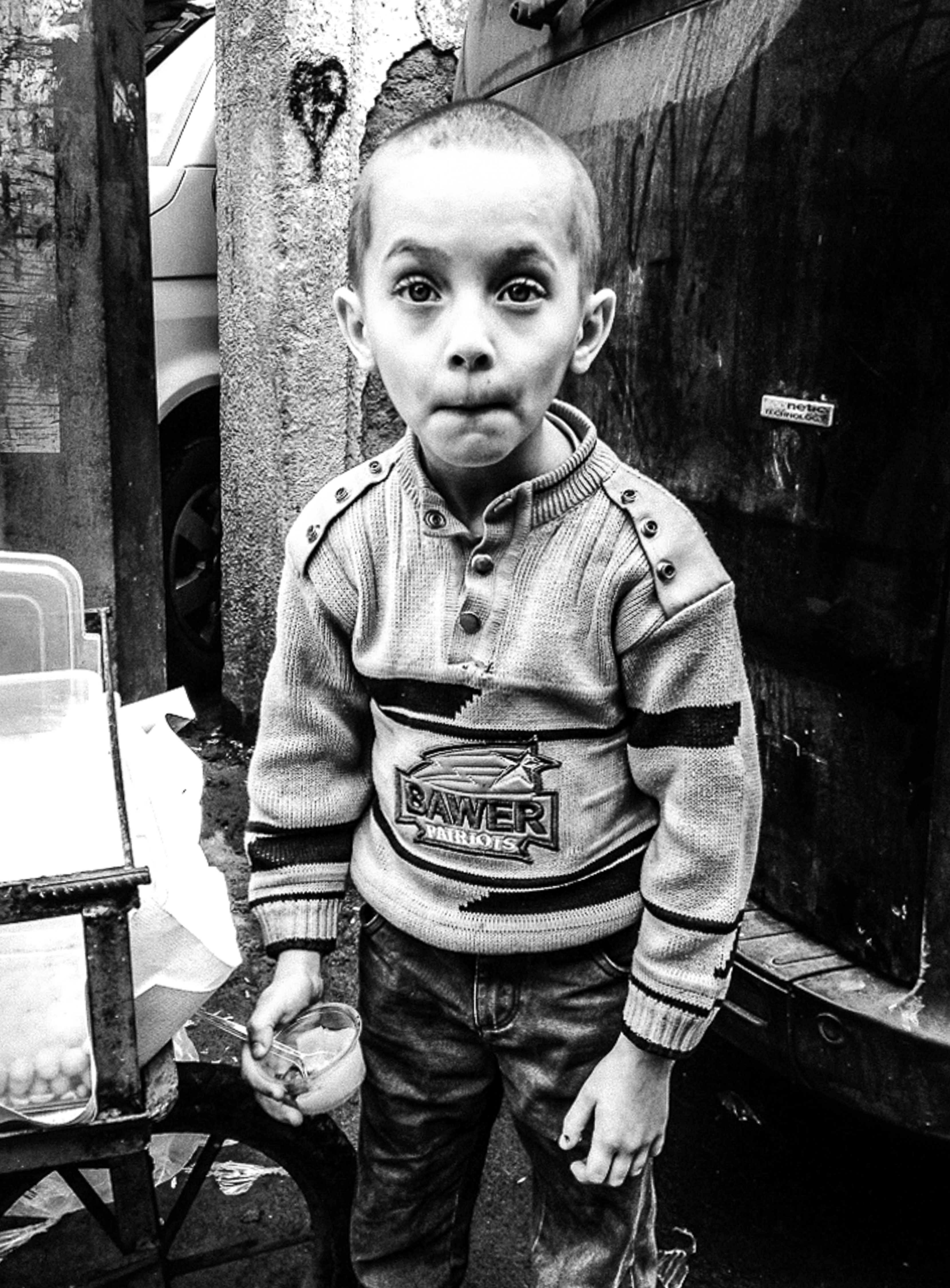 A young boy wearing a jumper with 'Sawer' logo, standing in a doorway against a graffitied wall.