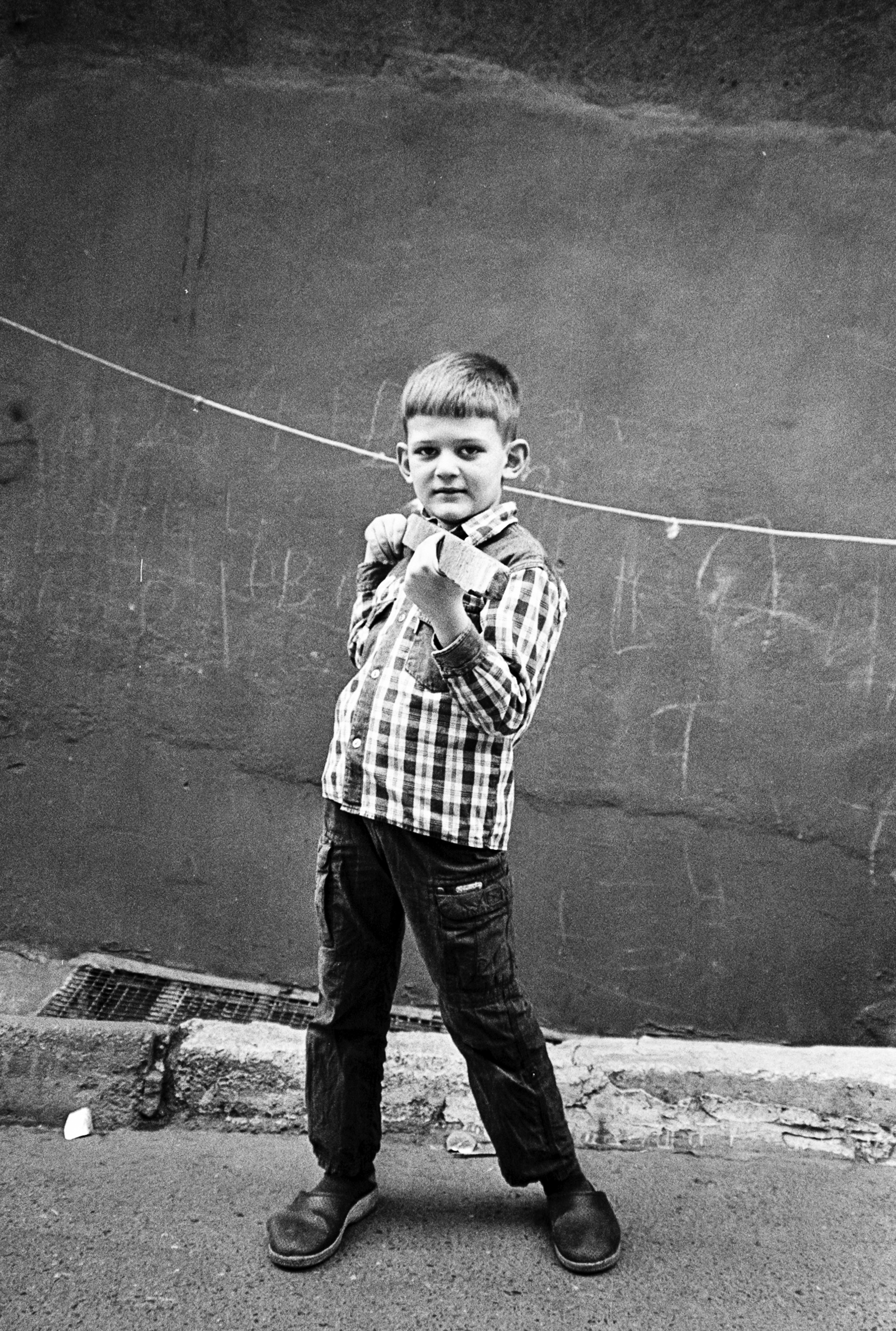 Young boy in checkered shirt and jeans, standing in front of a dark wall.