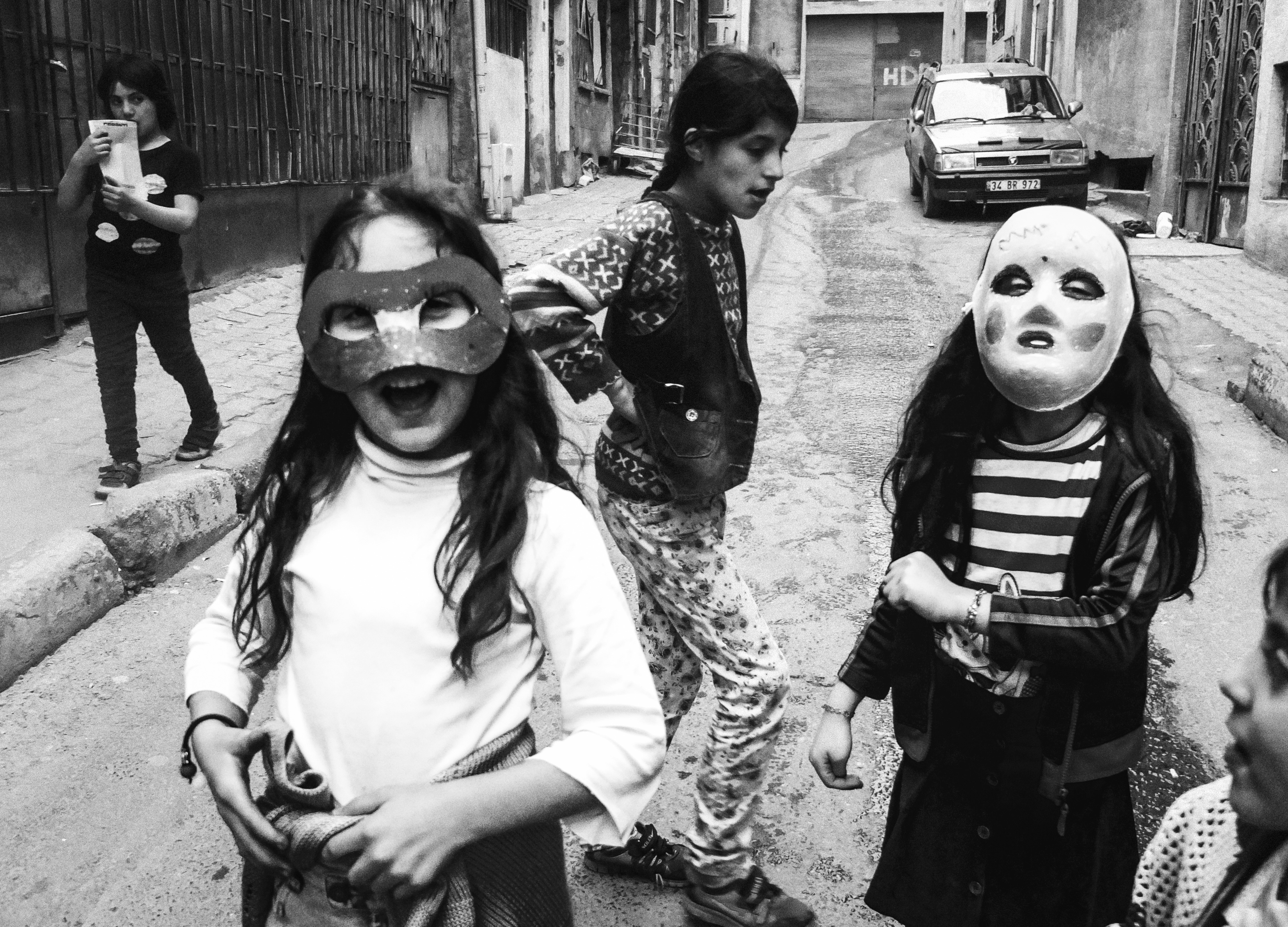 Three masked individuals walking down a narrow street, one wearing a leopard print dress and another in a striped top, with a car visible in the background.