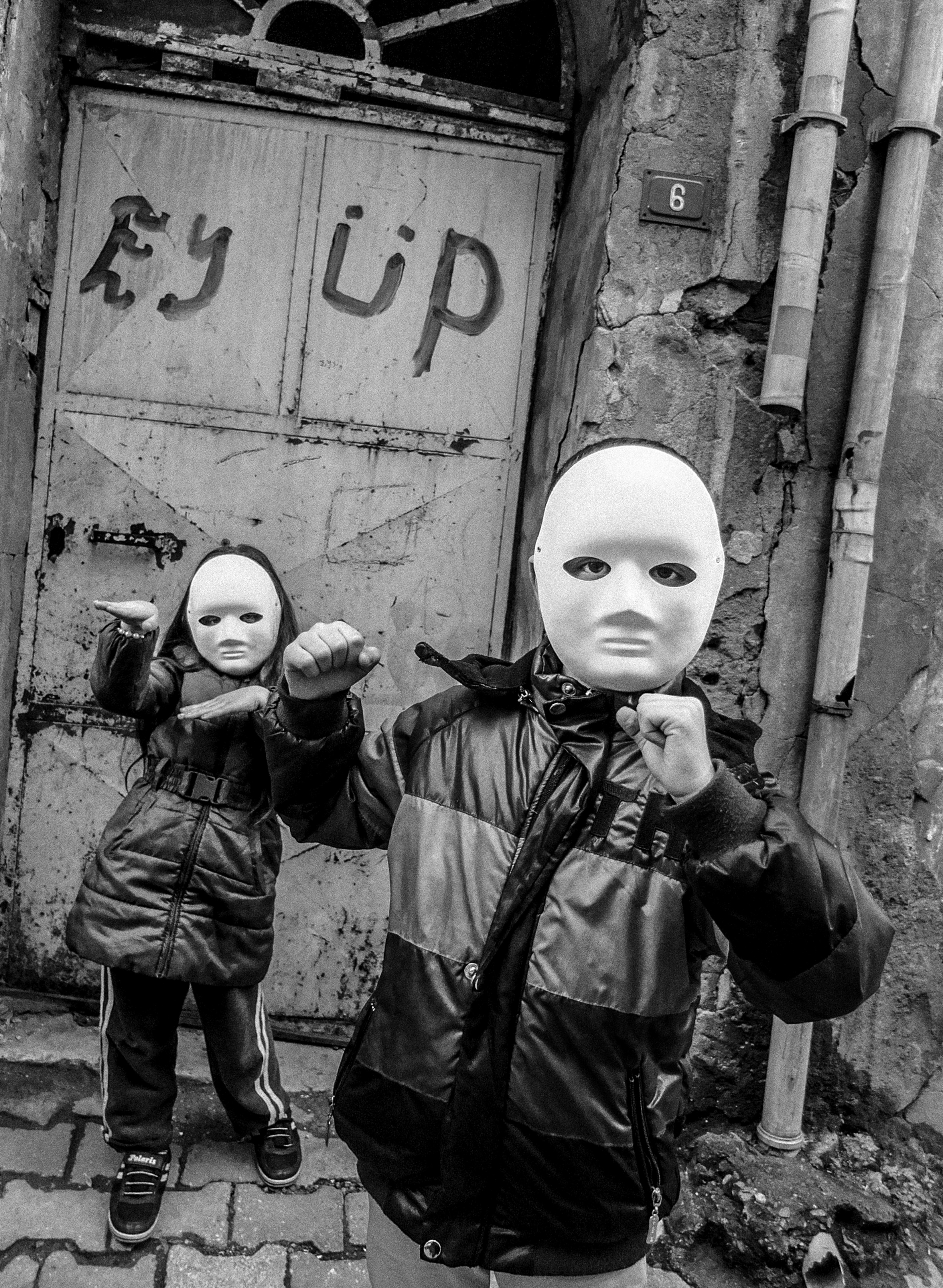 Two blank-faced figures in dark coats standing in front of a graffitied wall with the words "Ry up" visible.