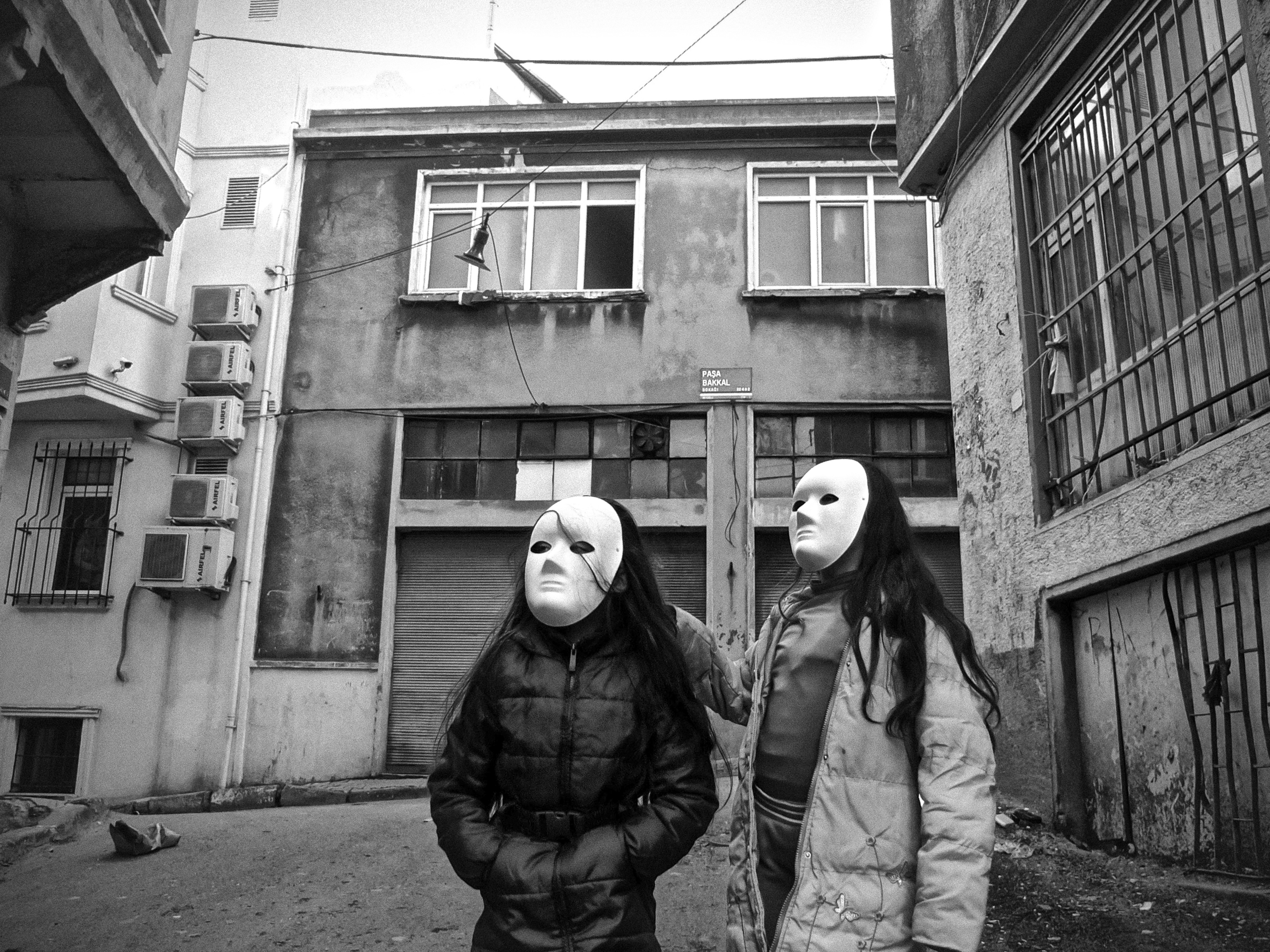 Monochrome image showing two individuals wearing masks, standing in front of a dilapidated building.