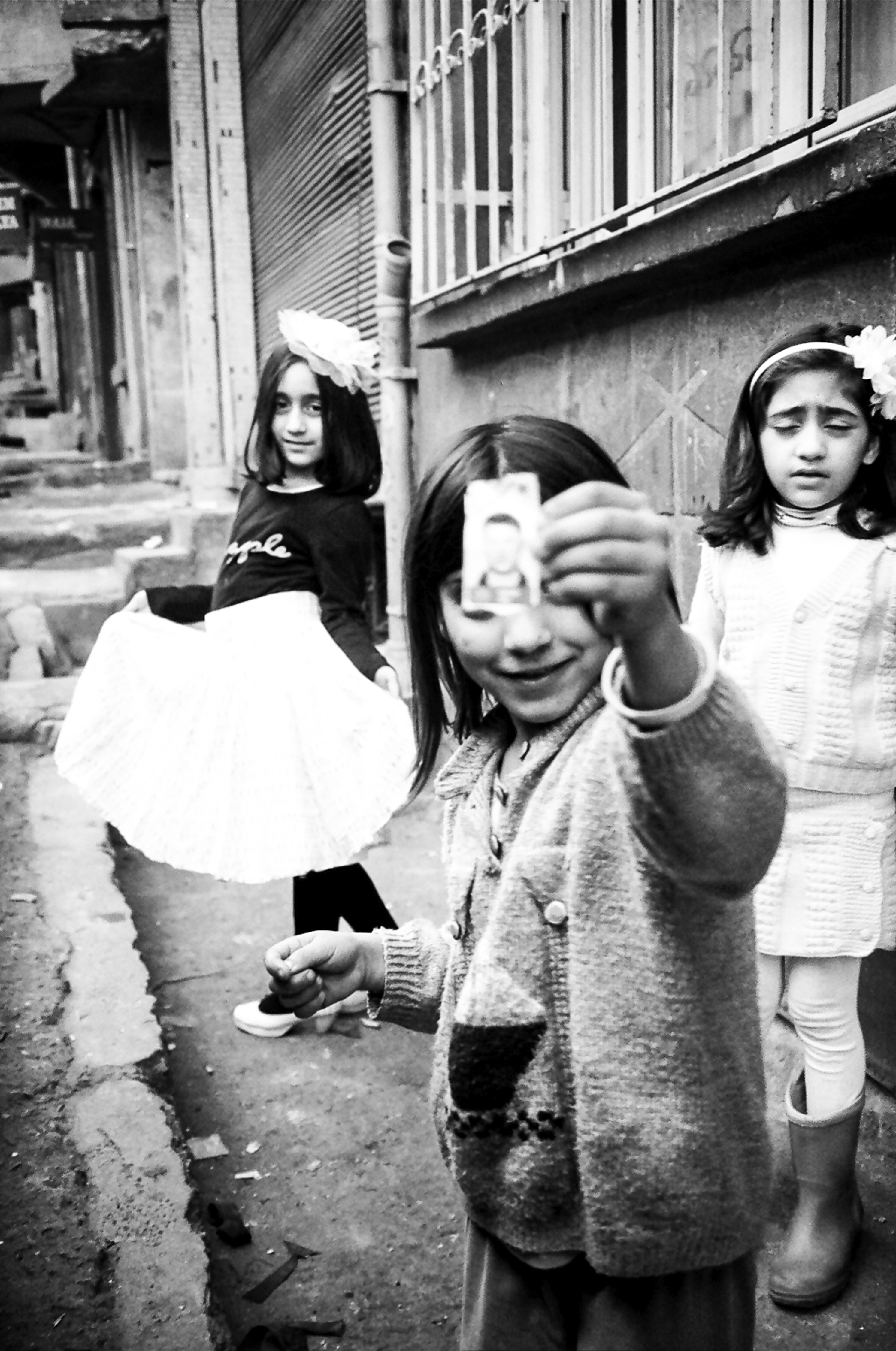 Three young girls, one taking a photo, in a black and white street scene with buildings in the background.