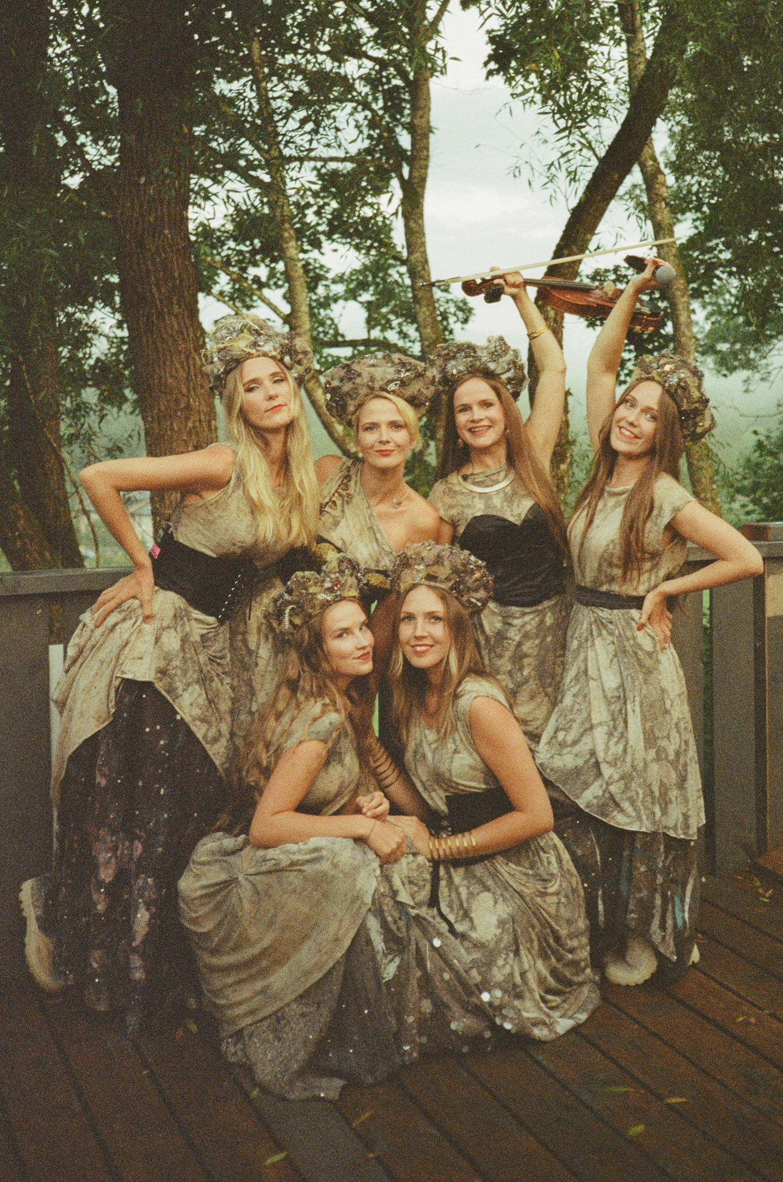 Group of females in elaborate floral-patterned dresses, posing together on a wooden deck surrounded by trees.