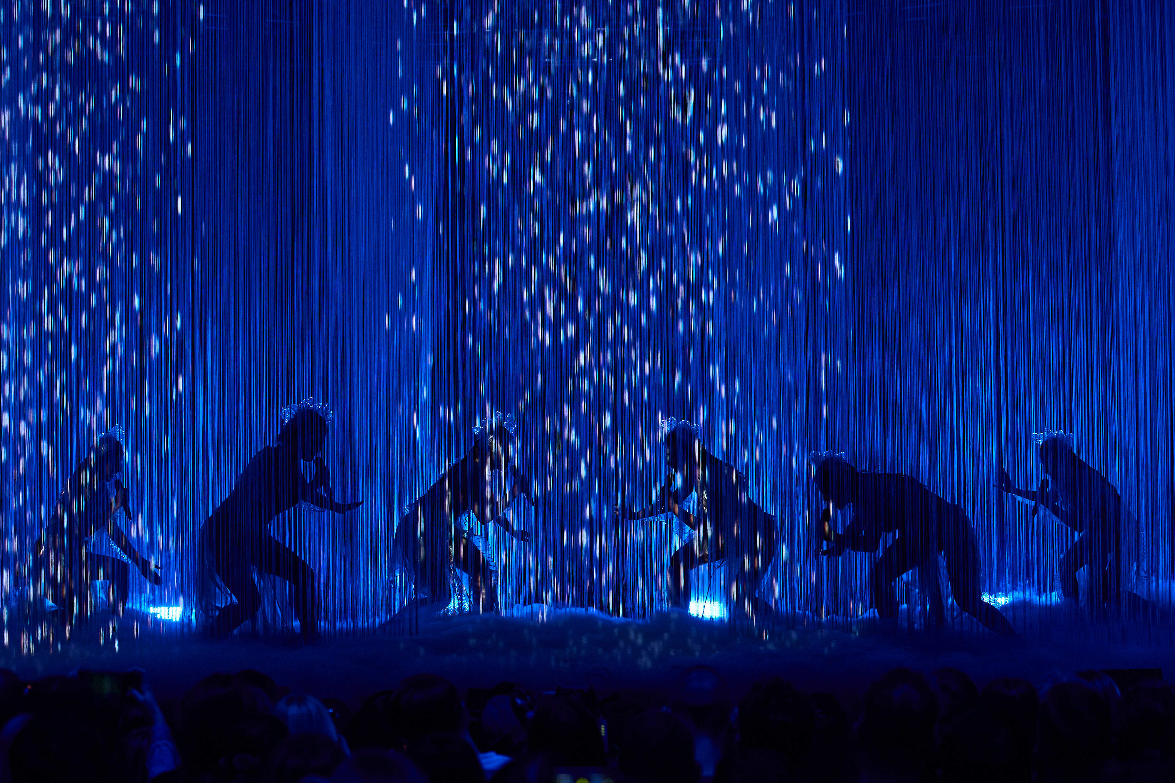 Blue rain-like lights falling on a stage with silhouettes of dancers performing.