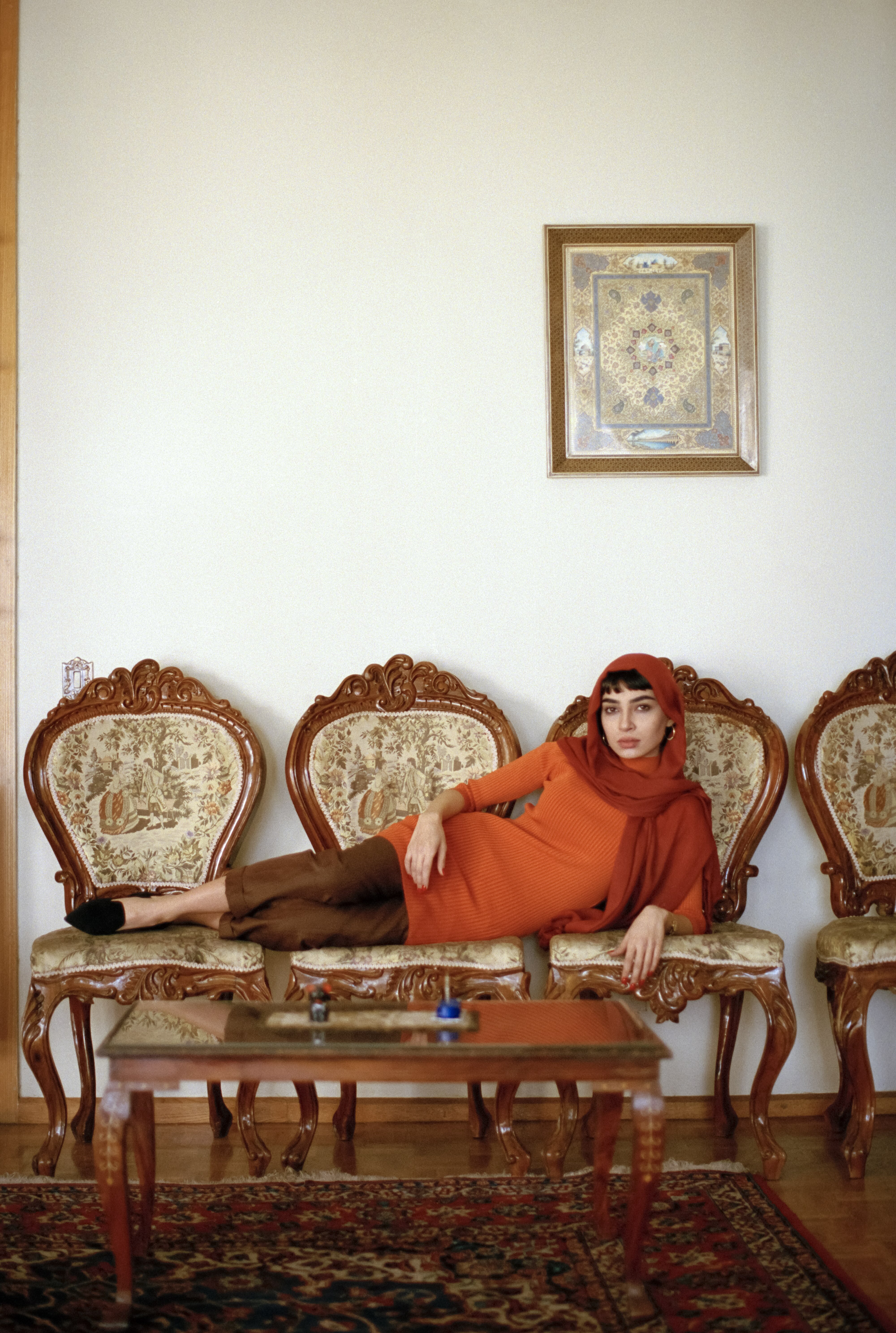 Ornate wooden chairs with floral upholstery, a patterned rug, and a framed artwork on the wall. A person in a vibrant orange outfit seated on the chairs.