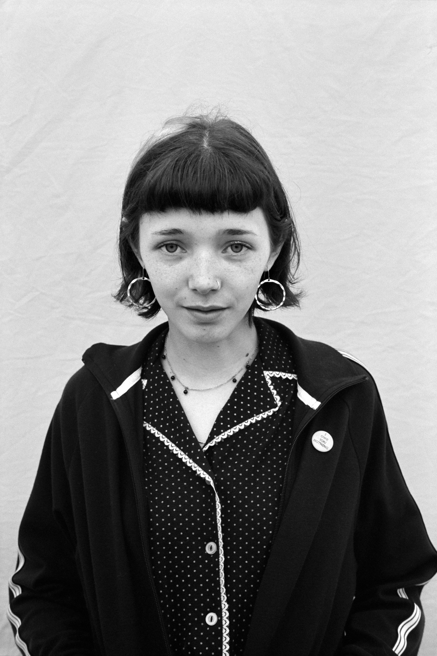 Black and white portrait of young woman with blunt bob haircut, wearing polka dot pyjama top and dark hoodie with circular earrings.
