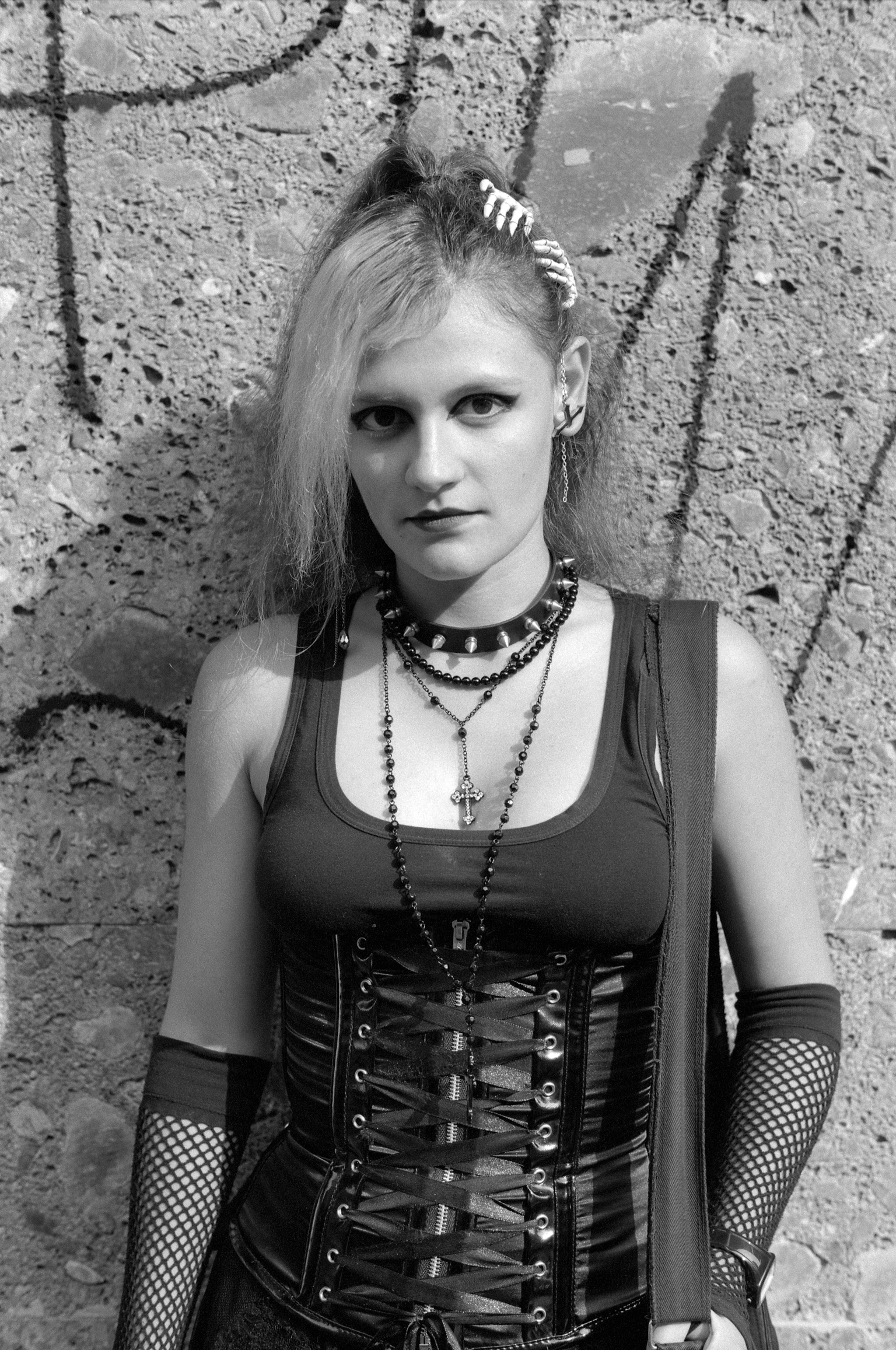 Woman in gothic attire with leather corset, fishnet sleeves, layered necklaces, and hair clips against textured stone wall background.