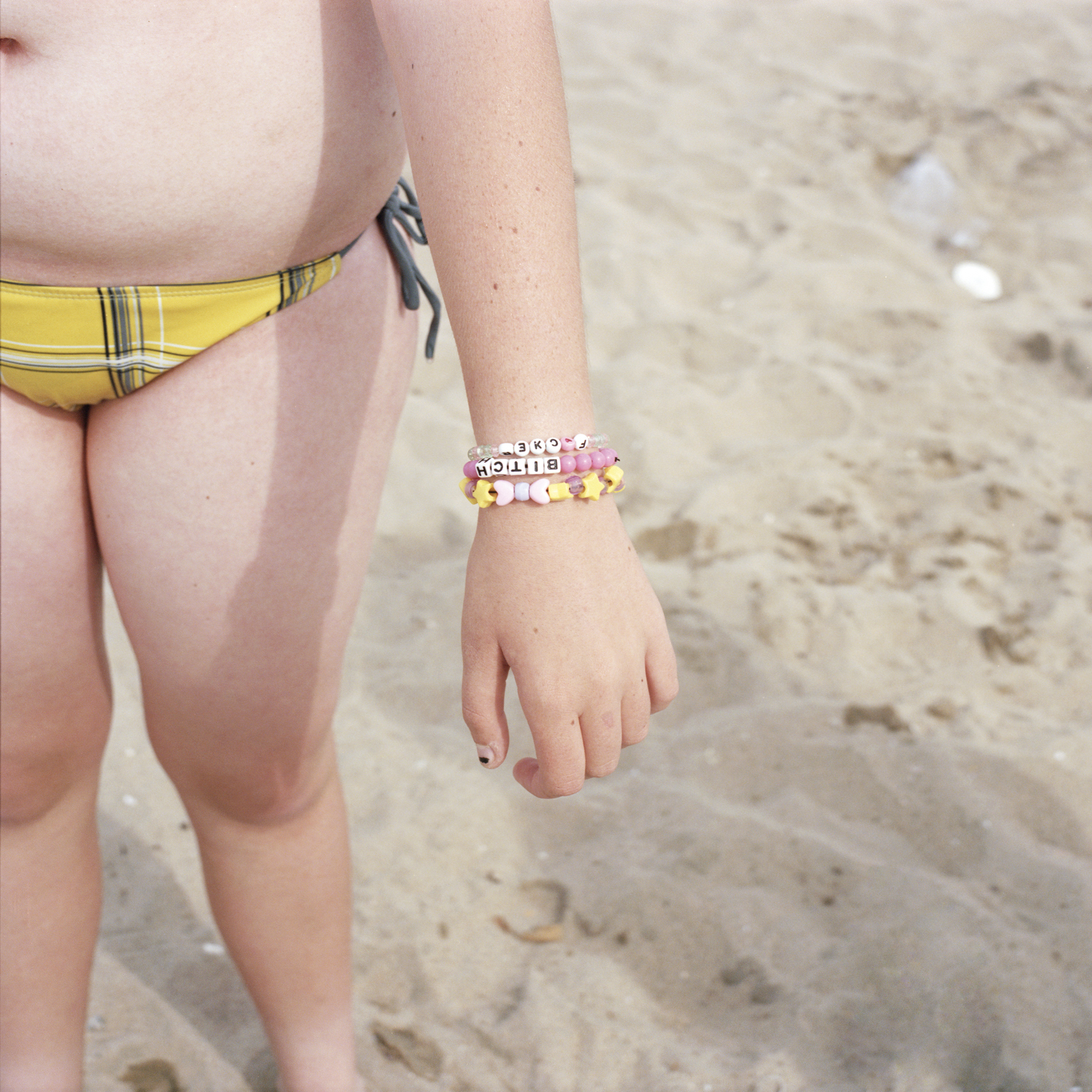Child wearing yellow bikini with colourful wristband holding snorkel gear, standing on sandy beach with footprints visible.