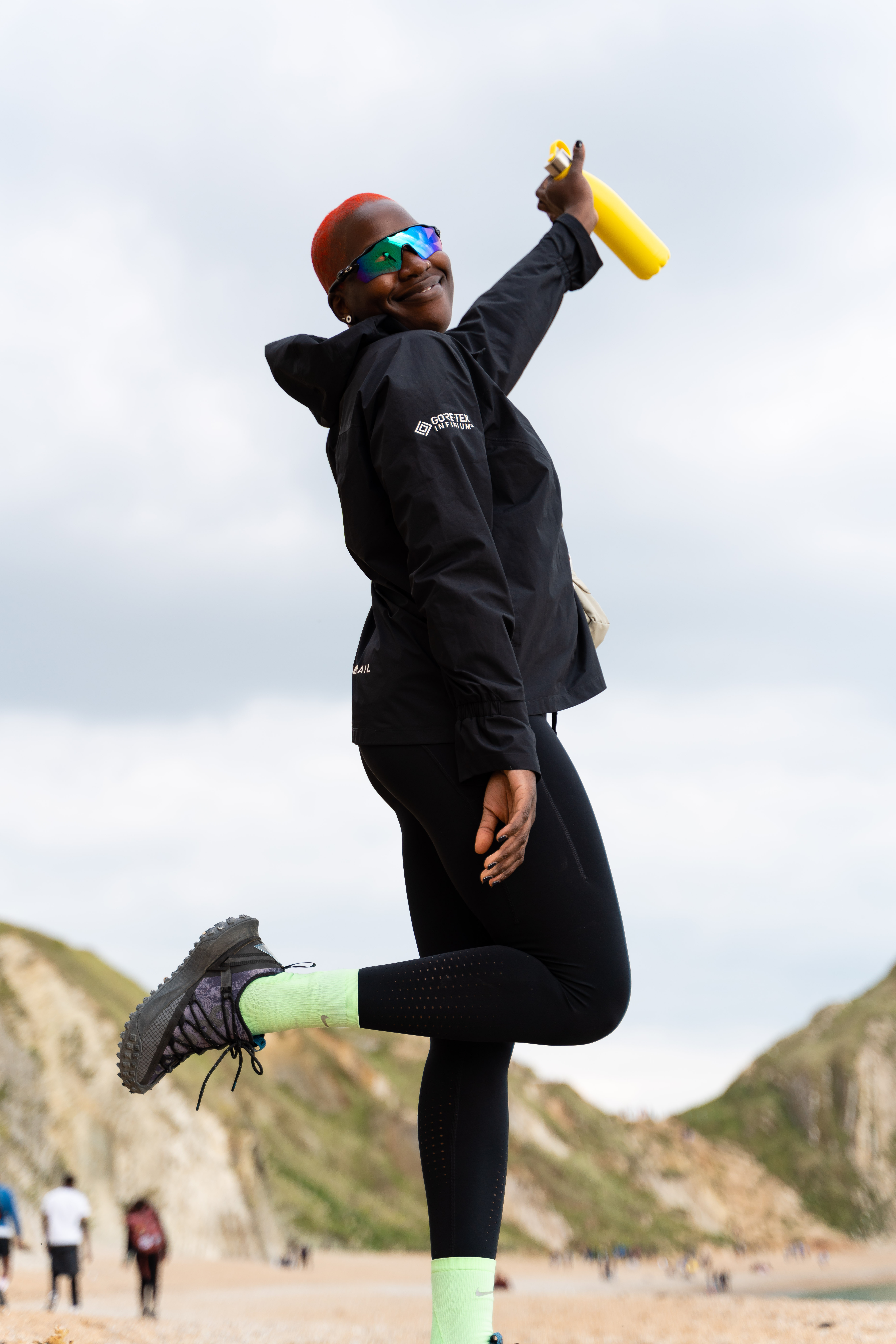 Person in black wetsuit and red helmet jumping on beach, holding yellow water bottle, with cliffs in background.